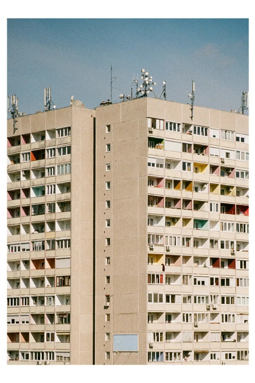 The Poster Club Block O’Clock, an urban photography print featuring a striking brutalist concrete apartment building with a repetitive grid of windows and balconies in muted pastel colors like pink, red, yellow, and blue.