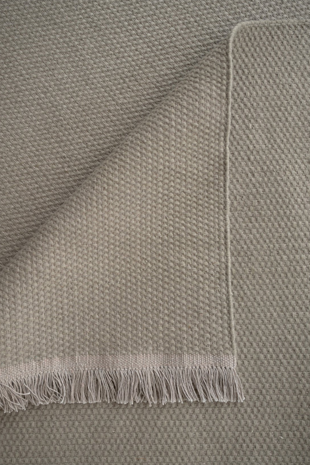 Linie Design Pastoral Unity Wool Rug, a premium hand-knotted wool area rug in a versatile greige neutral tone with a distinctive textured weave, offering a soft underfoot feel. This cozy rug is finished with delicate fringe detailing along one edge, making it ideal for Scandinavian living rooms, organic modern bedrooms, or contemporary minimalist spaces.
