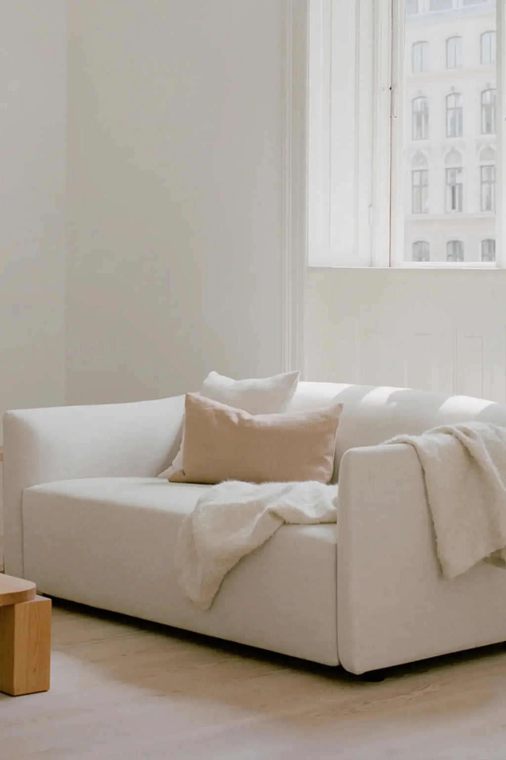 New Works Shore Sofa 2-Seater - Extended Base, a modern minimalist 2-seater sofa upholstered in a soft, textured cream fabric, featuring a low-profile silhouette, wide block arms, and plush deep seating for ultimate comfort. This Scandinavian-inspired sofa is perfectly accented with a light beige cushion and a cozy cream throw, making it an ideal choice for contemporary living rooms, minimalist apartments, or inviting lounge areas.