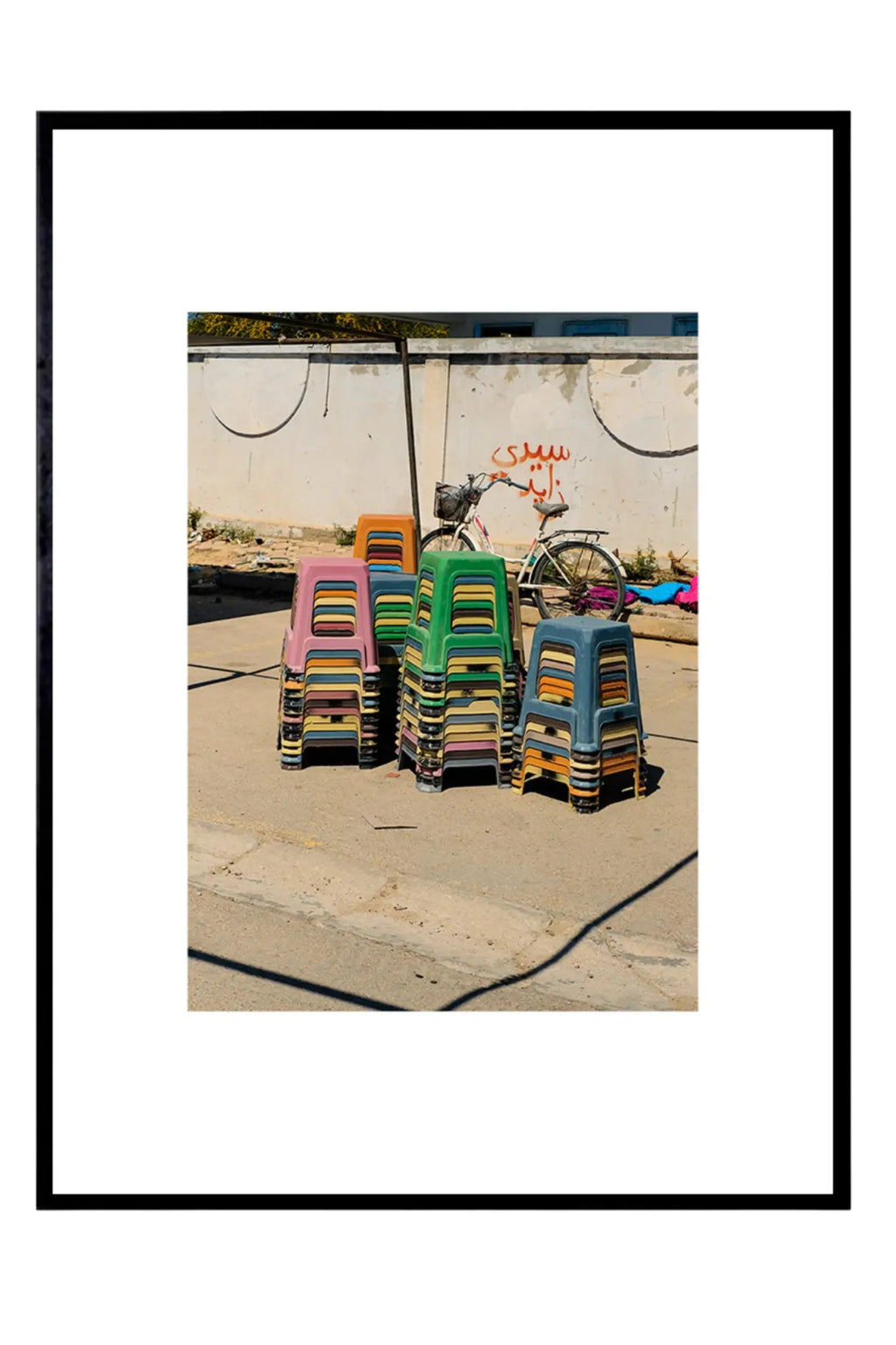 The Poster Club The Souk, a vibrant street photography art print featuring stacks of colorful plastic stools in shades of pink, green, blue, orange, and yellow, set against a textured beige wall with red graffiti and a bicycle. This contemporary urban art poster is presented in a sleek black frame with a white mat. Ideal for adding a pop of color and modern wall art appeal to contemporary living rooms, creative workspaces, or a dynamic gallery wall.