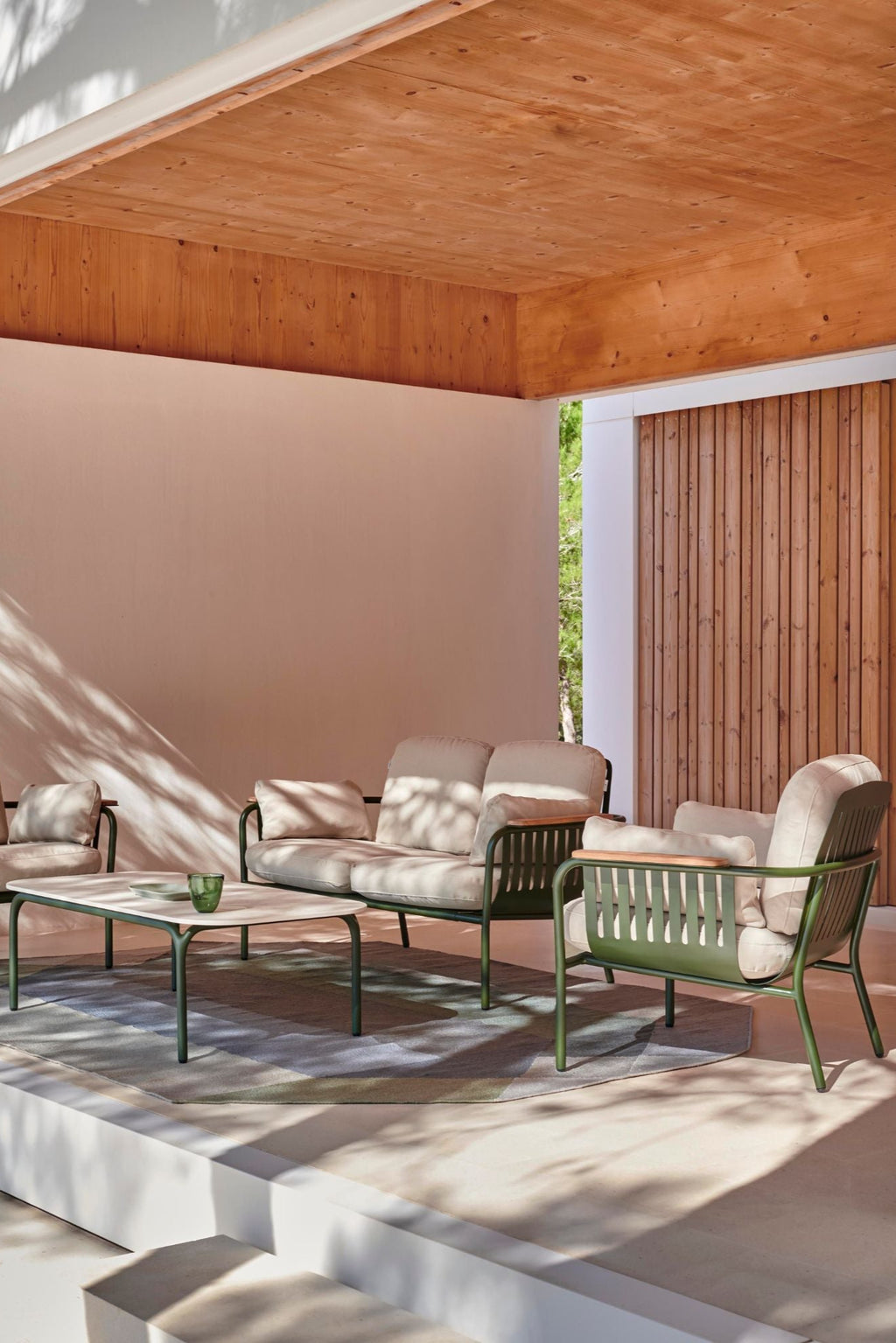 Gandia Blasco Capa 2-Seat Sofa, a modern minimalist outdoor sofa featuring a durable matte olive green aluminum frame with a slatted backrest and sleek curved legs, accented by natural wood armrests. It is upholstered with plush, light beige outdoor fabric cushions for ultimate comfort. This contemporary outdoor lounge sofa is perfect for stylish patio, deck, or garden living spaces.