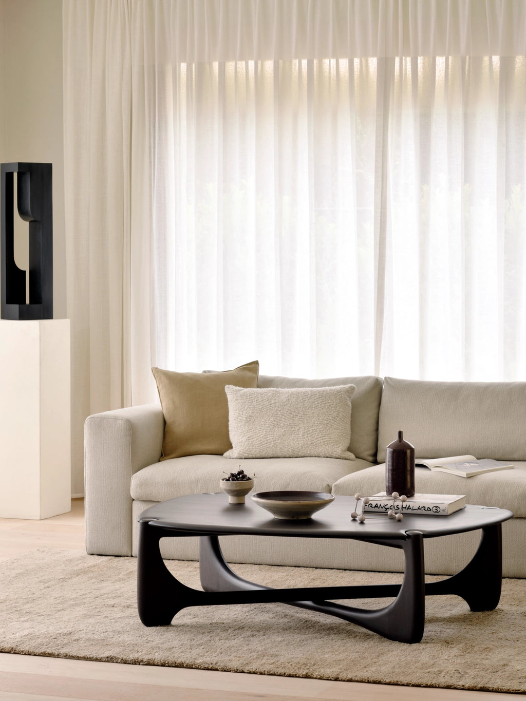 Ethnicraft PI Coffee Table, a dark brown oval wooden coffee table with elegantly curved legs, serving as a focal point in a stylish living room with a cream sofa and sheer white curtains.