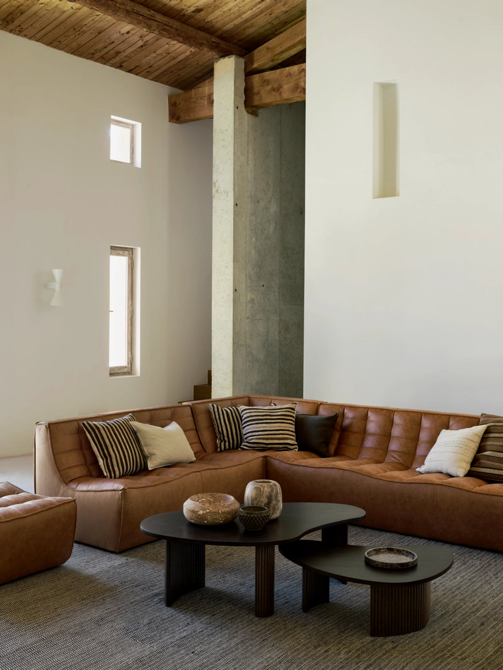 Ethnicraft Boomerang Coffee Table set, two dark brown mahogany tables with organic, curved shapes and textured slatted bases, anchoring a spacious living room with a large brown leather sectional sofa.