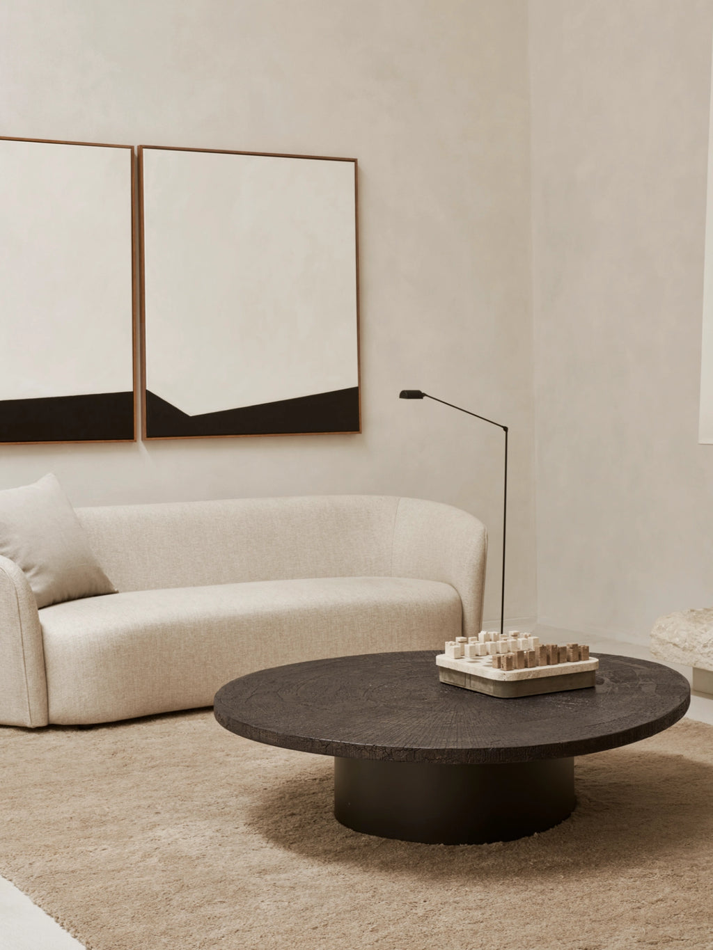 Ethnicraft Slice Coffee Table, an elegant oval-shaped table with a dark, richly textured surface and a solid, cylindrical base. It sits on a light beige rug in a minimalist living room, adorned with a stylish wooden chess set.