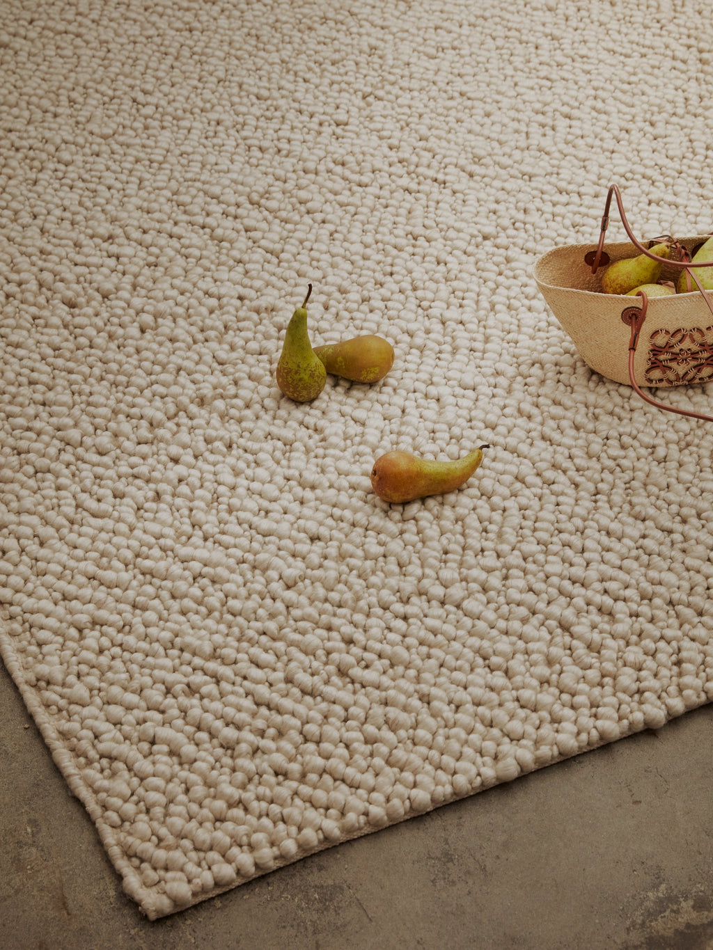 Tinted Objects Andersdotter Wool Rug, a plush, textured off-white natural wool area rug featuring a distinct looped or bobble design, laid on a concrete floor with scattered pears and a straw basket. This soft, durable rug is perfect for adding warmth and sophisticated style to modern living spaces or bedrooms.