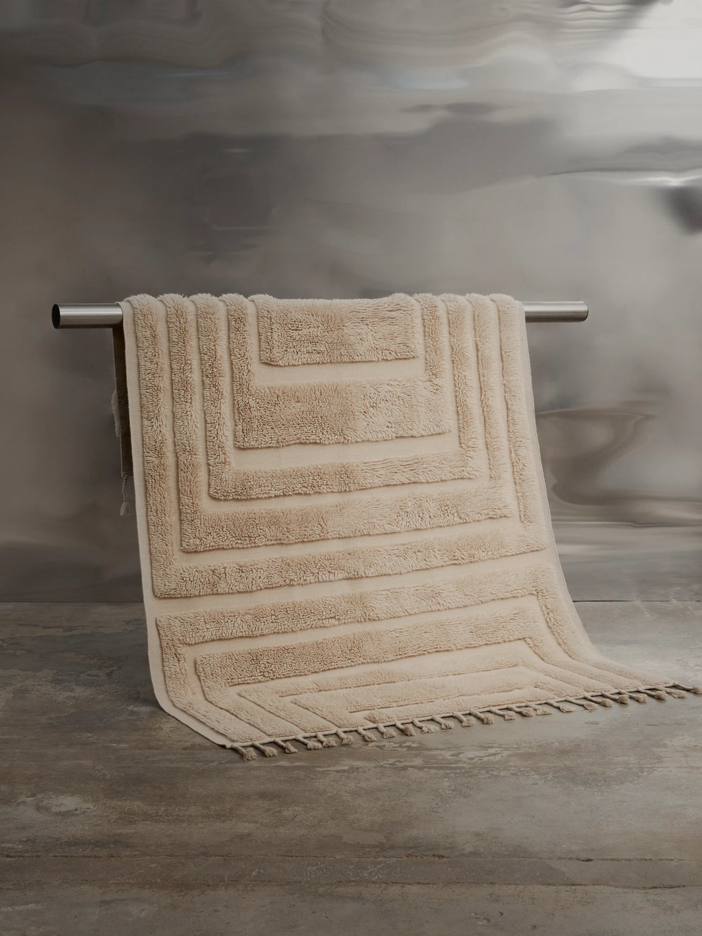 Tinted Objects Kask Wool Rug, a plush, textured cream-beige wool rug with an elevated geometric maze pattern and decorative tassels, draped over a silver bar. This stylish and soft area rug is perfect for adding warmth and modern design to any living space or bedroom.