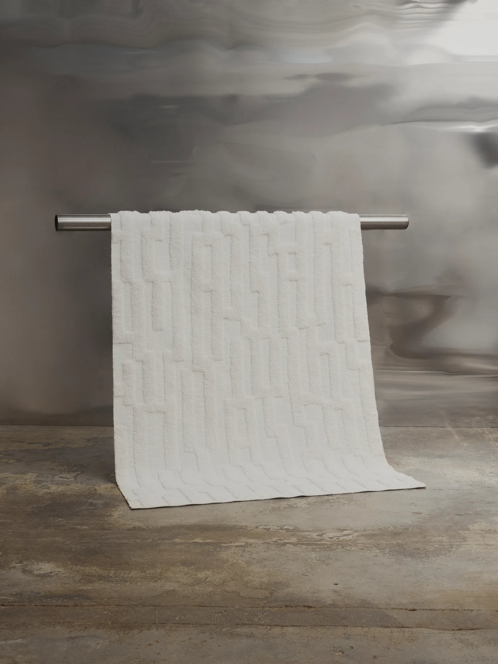 Tinted Objects Bielke Wool Rug, a plush, off-white rectangular rug with an elegant raised geometric pattern, draped over a silver rod on a concrete floor. This luxurious and soft area rug adds warmth and modern style to any living space or bedroom.