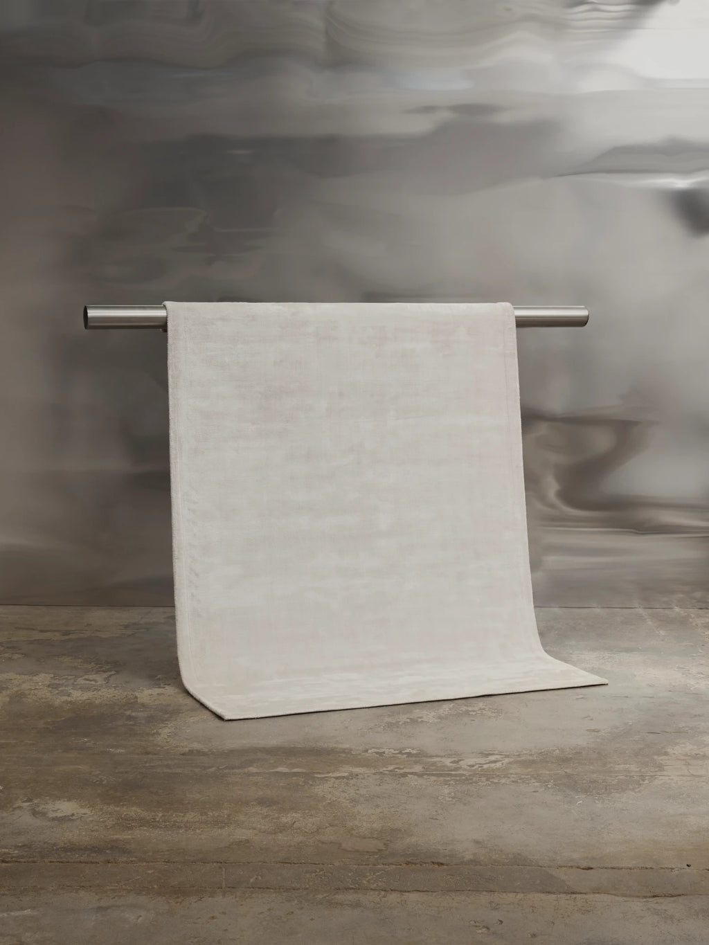 Tinted Objects Backfjall Viscose Rug, a luxurious light beige or off-white modern rug with a subtle sheen, draped over a silver rod against a mottled grey wall, unfurling onto a rough concrete floor. This elegant area rug is perfect for adding soft texture and contemporary style to a sophisticated living room or bedroom.