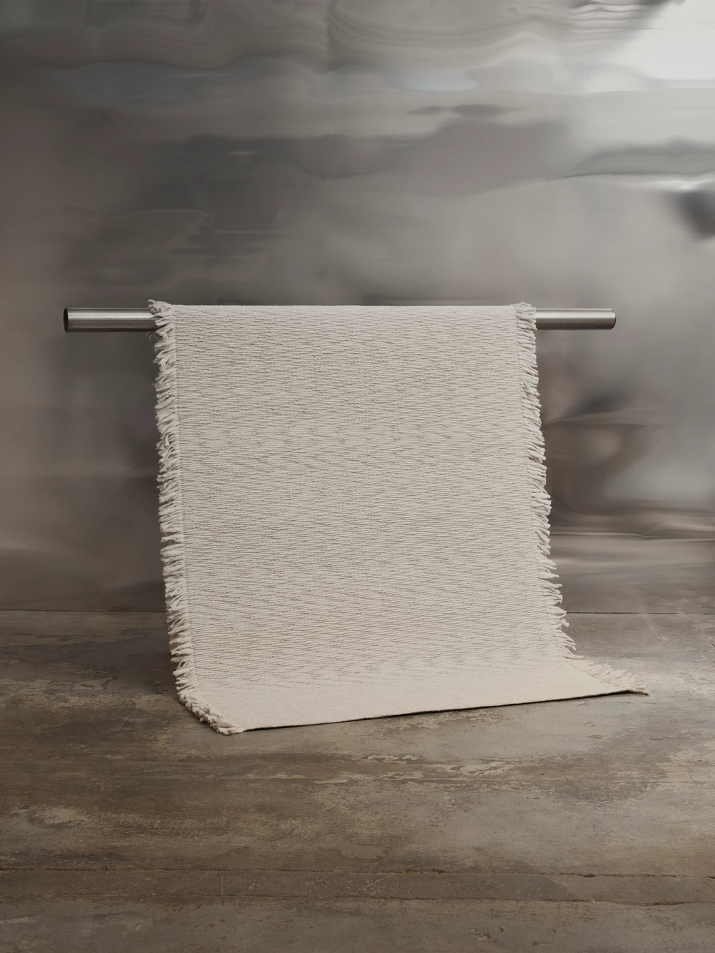 Tinted Objects Fagerlund Wool Rug, a plush off-white textured area rug with delicate fringe edges, draped over a metal bar against a metallic wall. This luxurious, soft wool rug is perfect for adding warmth and sophisticated style to any modern living space or bedroom.