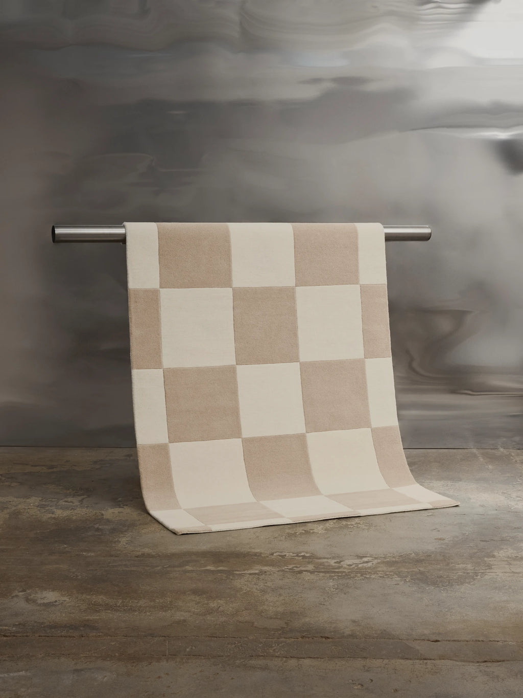 Tinted Objects Hafstrom Wool Rug, featuring a checkerboard pattern in soft beige and cream squares, draped over a polished metal bar. This stylish, high-quality wool rug adds warmth and modern elegance to any contemporary living space or bedroom.
