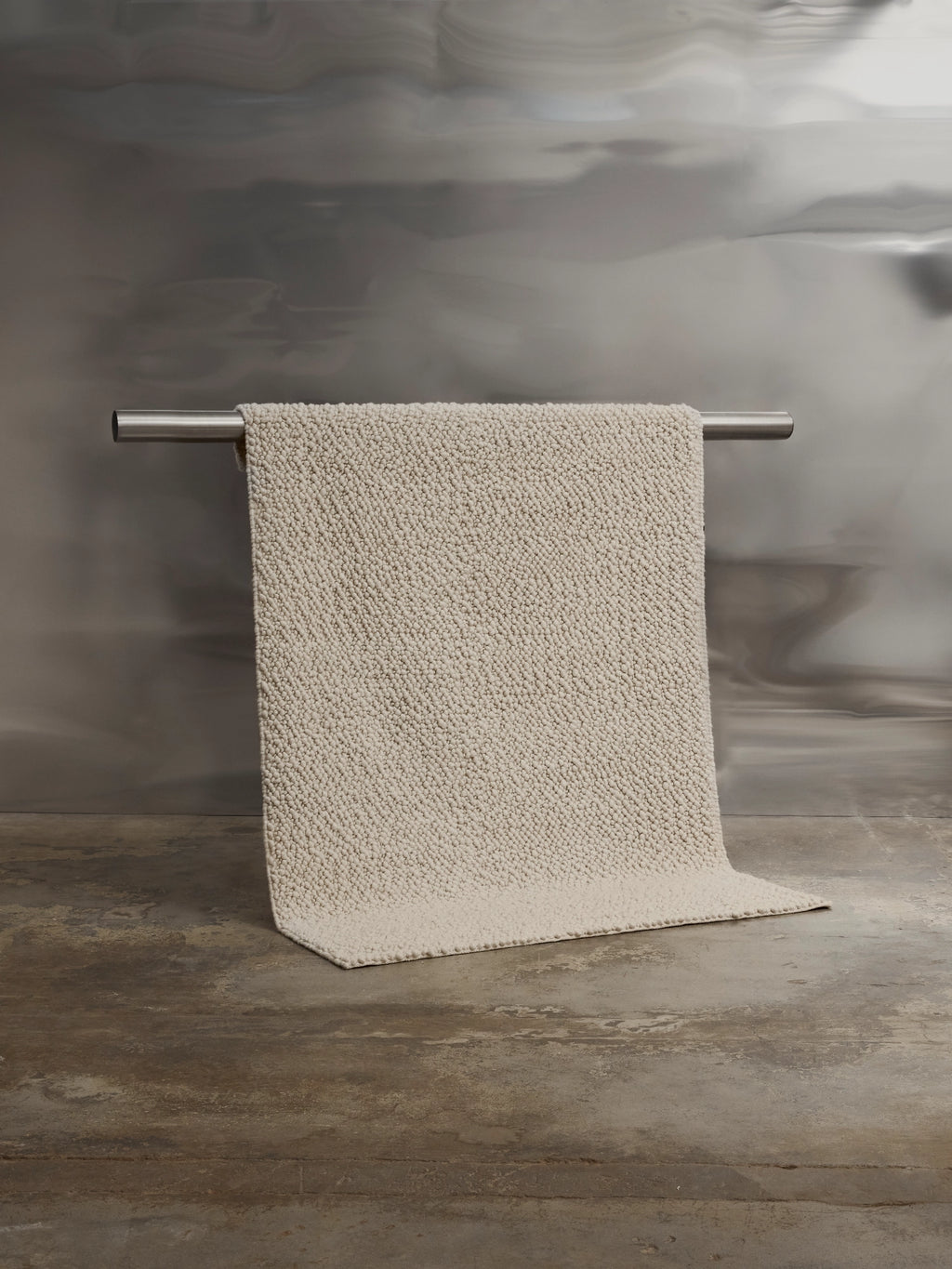 Tinted Objects Andersdotter Wool Rug, a luxurious cream-colored textured wool rug with a soft, nubby surface, draped over a sleek silver bar. This premium natural fiber rug adds warmth and modern elegance to any living space or bedroom.