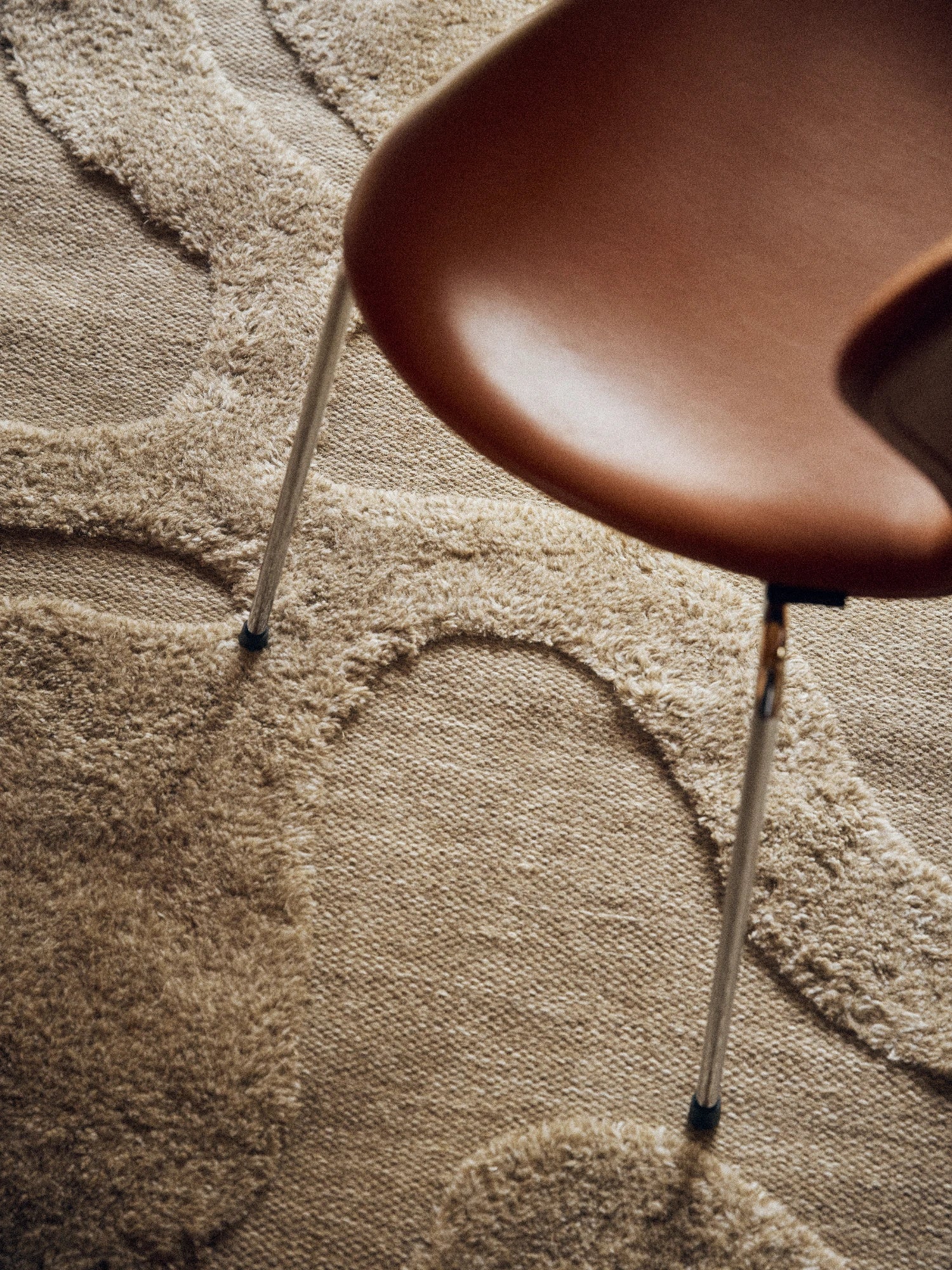 Tinted Objects Norlander Wool Rug, featuring a textured natural beige loop pile and cut pile geometric pattern, with a brown chair leg. This luxurious area rug adds warmth and modern style to any living space or bedroom.