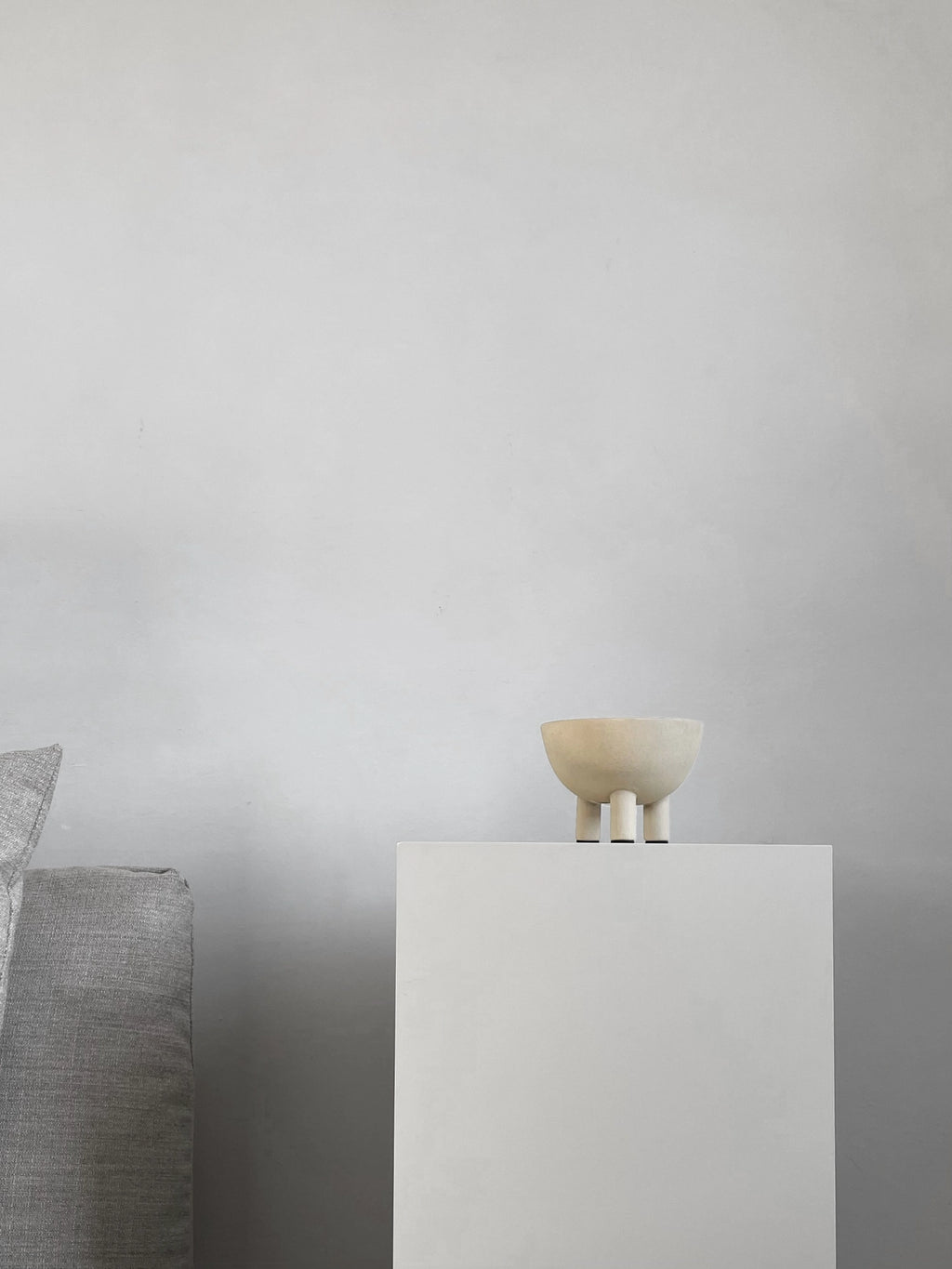 101 CPH Duck Bowl, a cream-colored, textured ceramic bowl with three short, dark-tipped legs, displayed on a minimalist white pedestal beside a gray fabric sofa in a studio setting. This premium home decor accent is ideal for modern interior styling.
