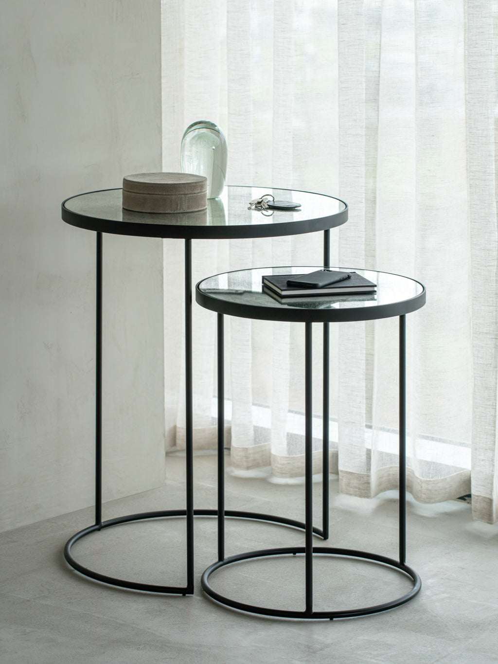 Ethnicraft Nesting Coffee Table Set with two clear glass-topped tables, featuring sleek black metal bases. The taller table holds a decorative box and keys, set against a window with sheer curtains.