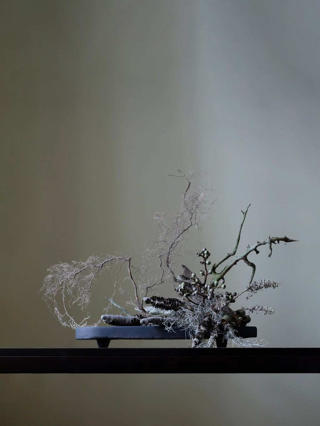 101 CPH Geta Tray, a low-profile, dark gray cement tray with short feet, displaying a natural arrangement of dried branches, seed pods, and white wispy elements. A premium decorative accent.
