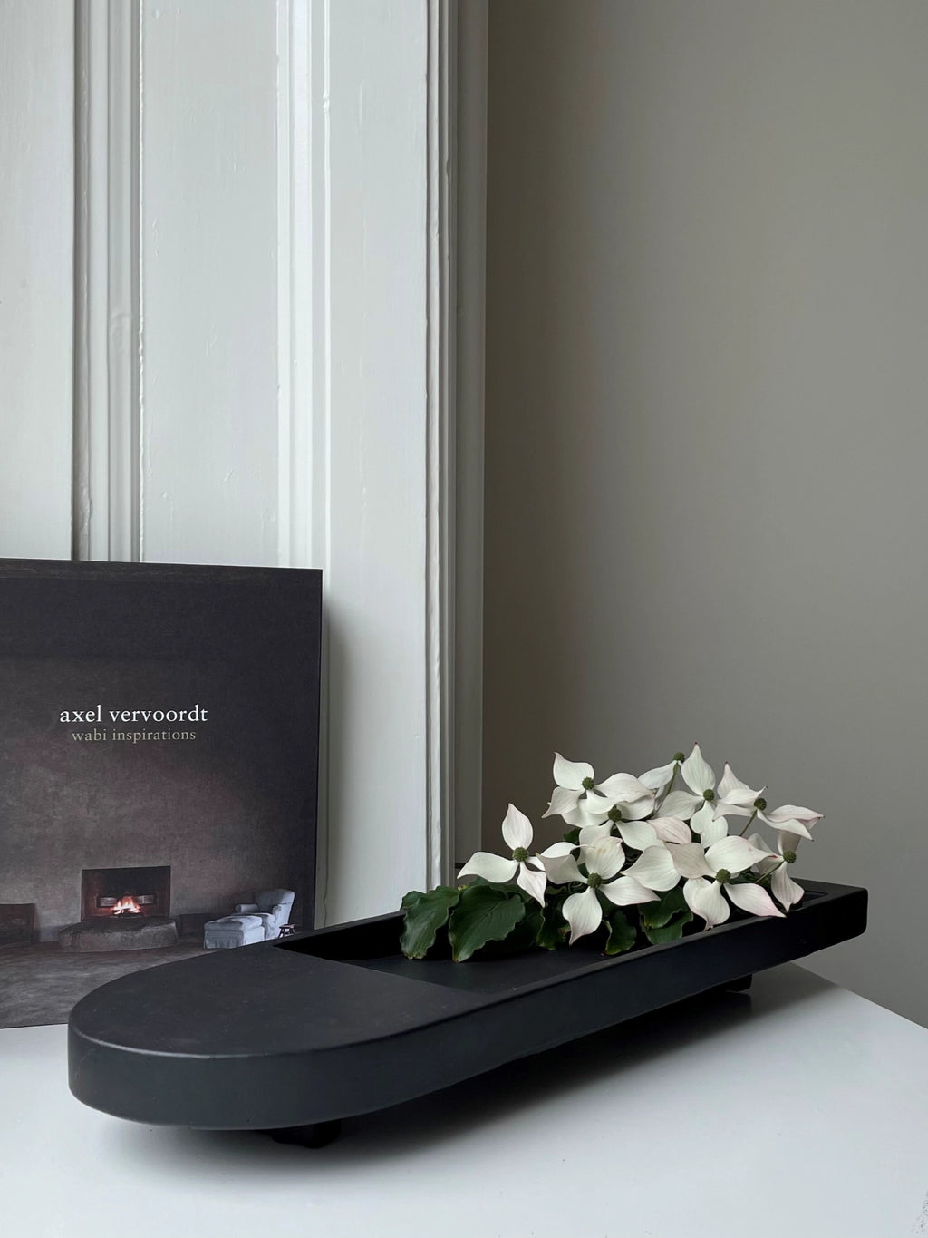 101 CPH Geta Tray, a modern black oblong ceramic tray with raised edges and small feet, holding white flowers on a white surface next to a book. This luxury decorative tray is perfect for sophisticated home accents.