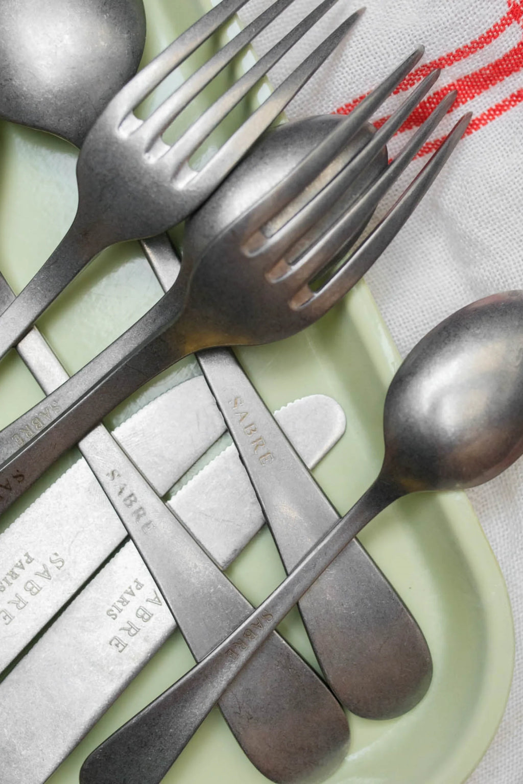 Sabre Paris Marius 5-Piece Cutlery Flatware Set, featuring rustic matte silver-toned stainless steel with visible "Sabre Paris" branding on each handle, arranged on a light green tray. This durable and stylish flatware set is perfect for everyday dining or special occasions for those who appreciate classic, understated elegance.