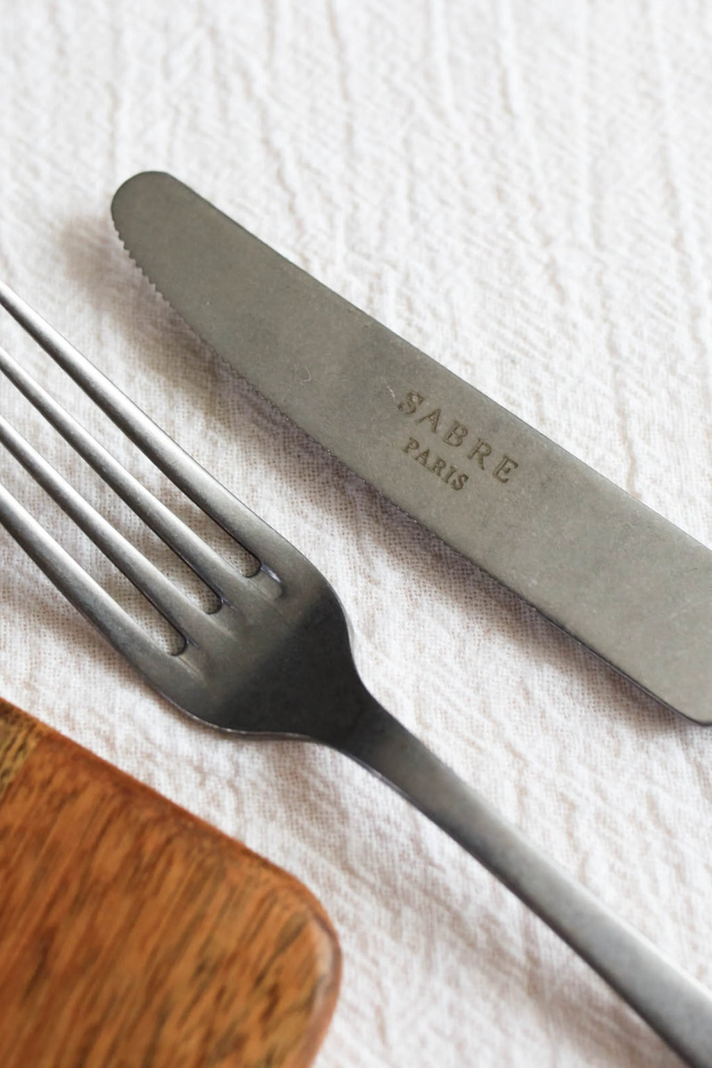 Sabre Paris Marius 5-Piece Cutlery Flatware Set, featuring a matte stainless steel knife with a serrated edge and a sleek four-tine fork, alongside a rich brown wooden handle. This elegant flatware is perfect for enhancing modern dining experiences for discerning hosts and home chefs.