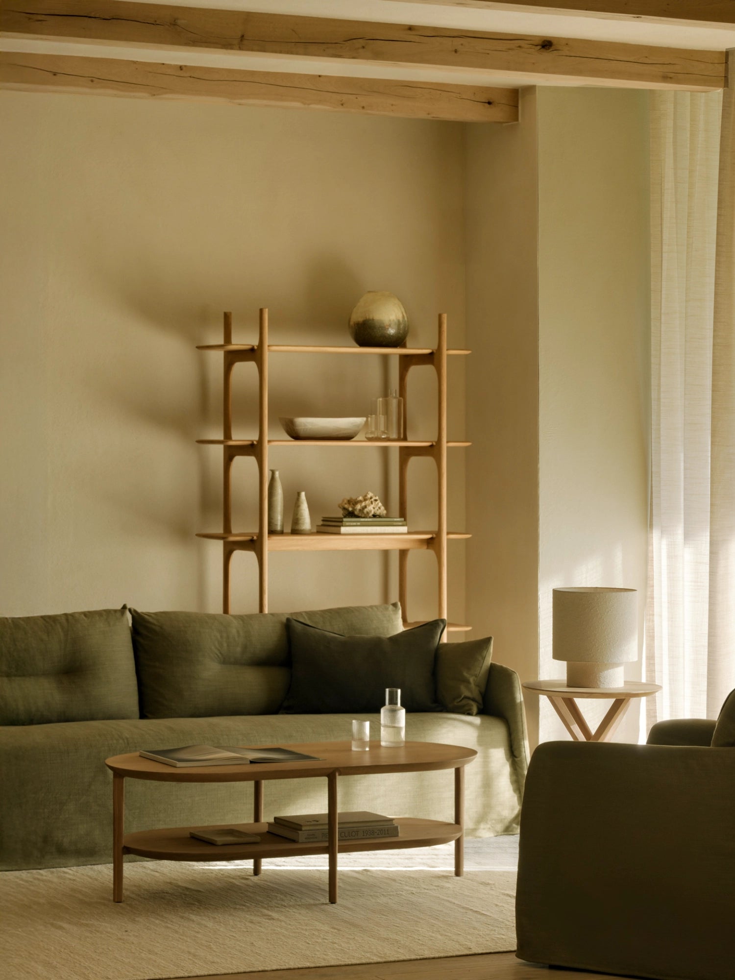 Ethnicraft Bok Coffee Table, a sleek, oval-shaped table crafted from light oak wood, featuring two tiers for storage. It sits on a neutral rug in a warm, inviting living room with a green linen sofa and wooden shelving unit.