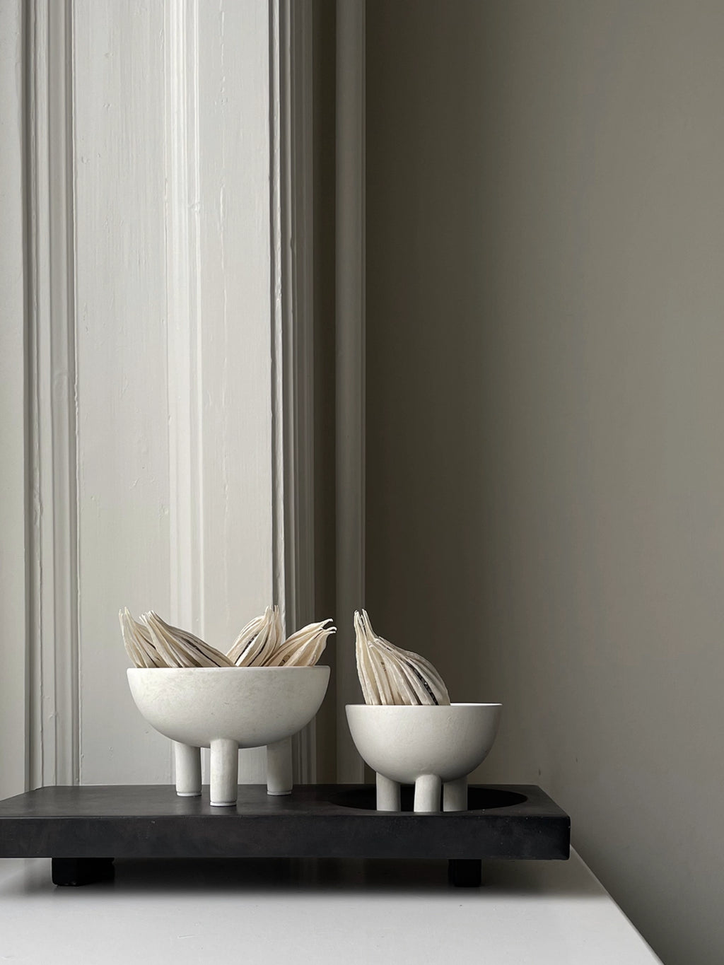 101 CPH Duck Bowl. Two off-white ceramic shallow bowls with short cylindrical legs, one larger and one smaller, sitting on a dark rectangle tray with dried botanical elements. A chic accent for modern home decor.