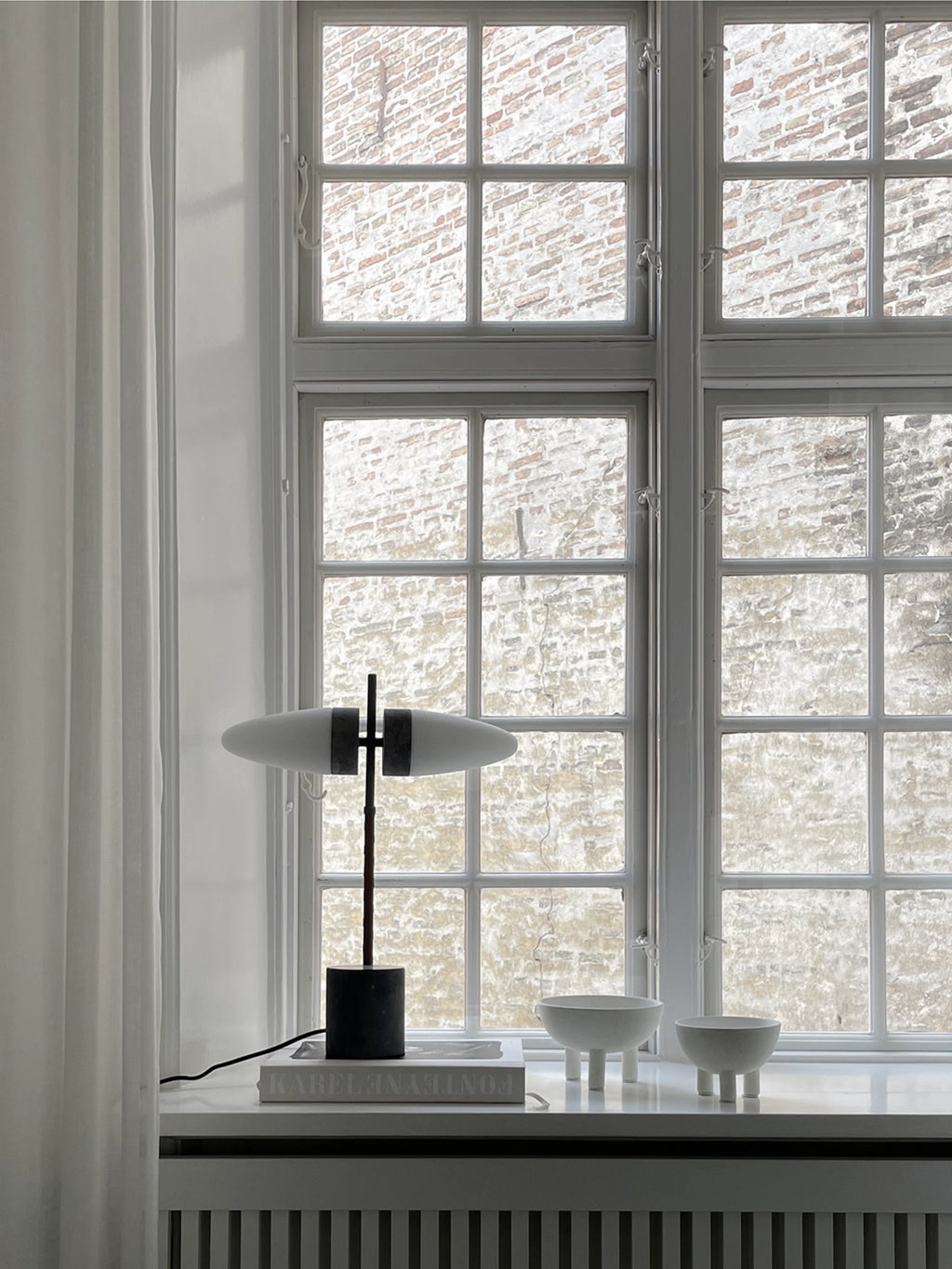 101 CPH Duck Bowl; Two matte white ceramic bowls with three sturdy legs each, displayed on a windowsill next to a lamp and books. Unique, minimalist homeware addition.