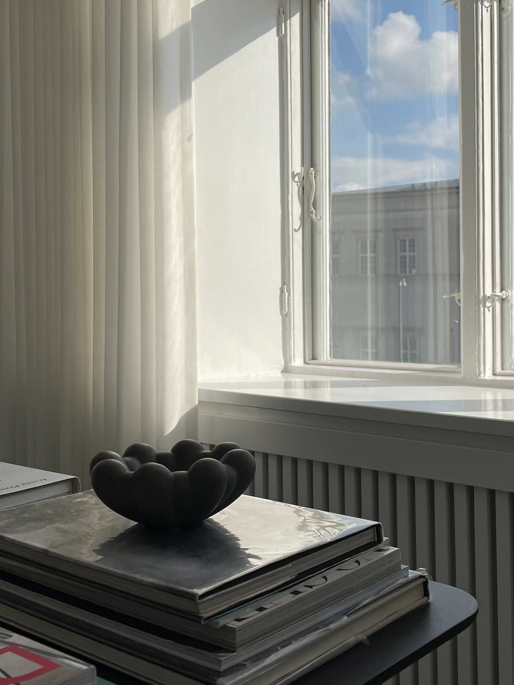 101 CPH Bloom Tray in a deep charcoal black, organic, petal-like shape, crafted from premium ceramic. Displayed on a stack of books by a bright window. Ideal for modern minimalist decor.