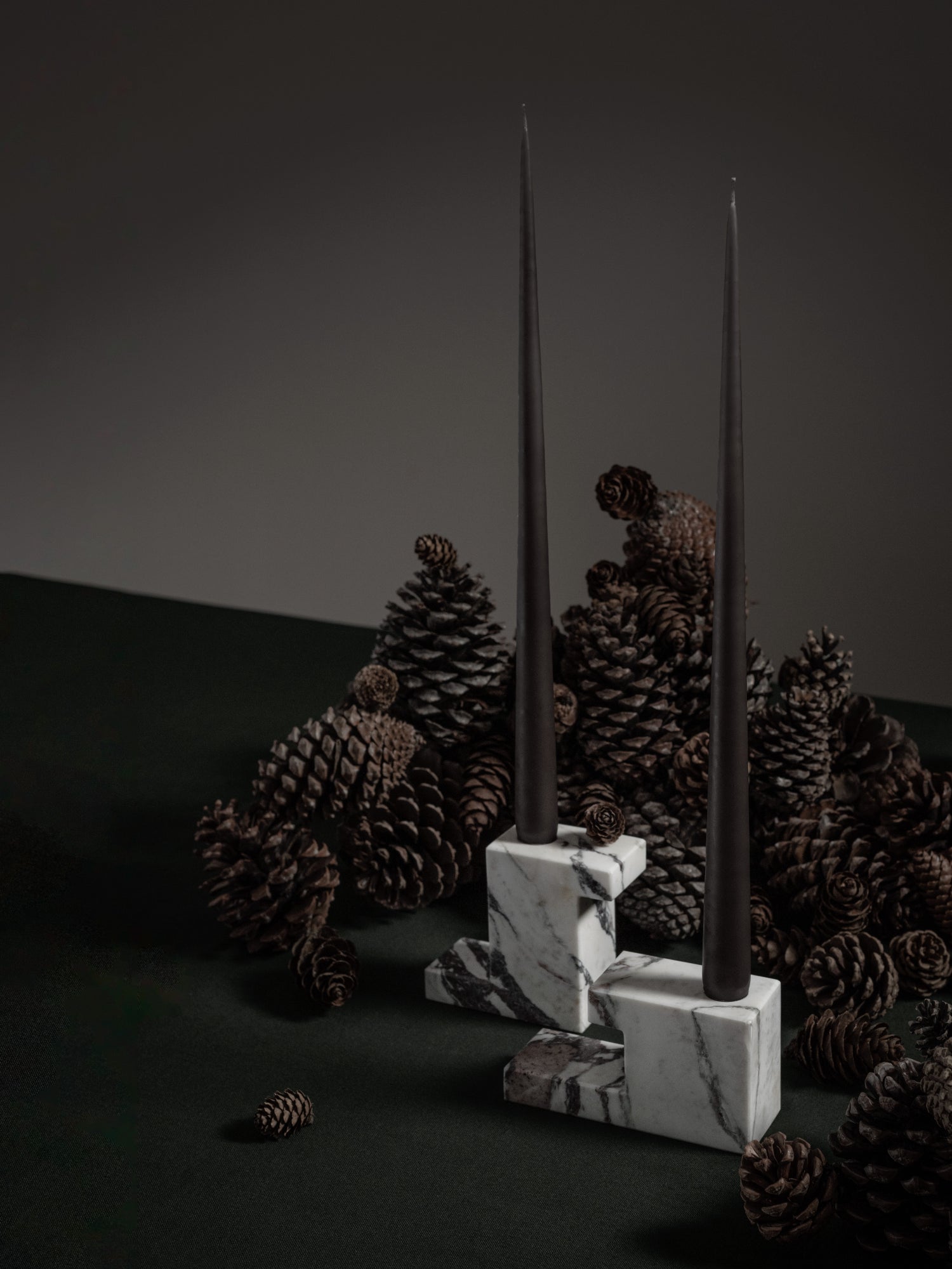 101 CPH Brick Candle Holder - Calacatta, a premium, modern square white Calacatta marble design with elegant grey veining, holding two tall black taper candles, styled amidst pinecones on a dark green surface. Ideal for sophisticated home decor.
