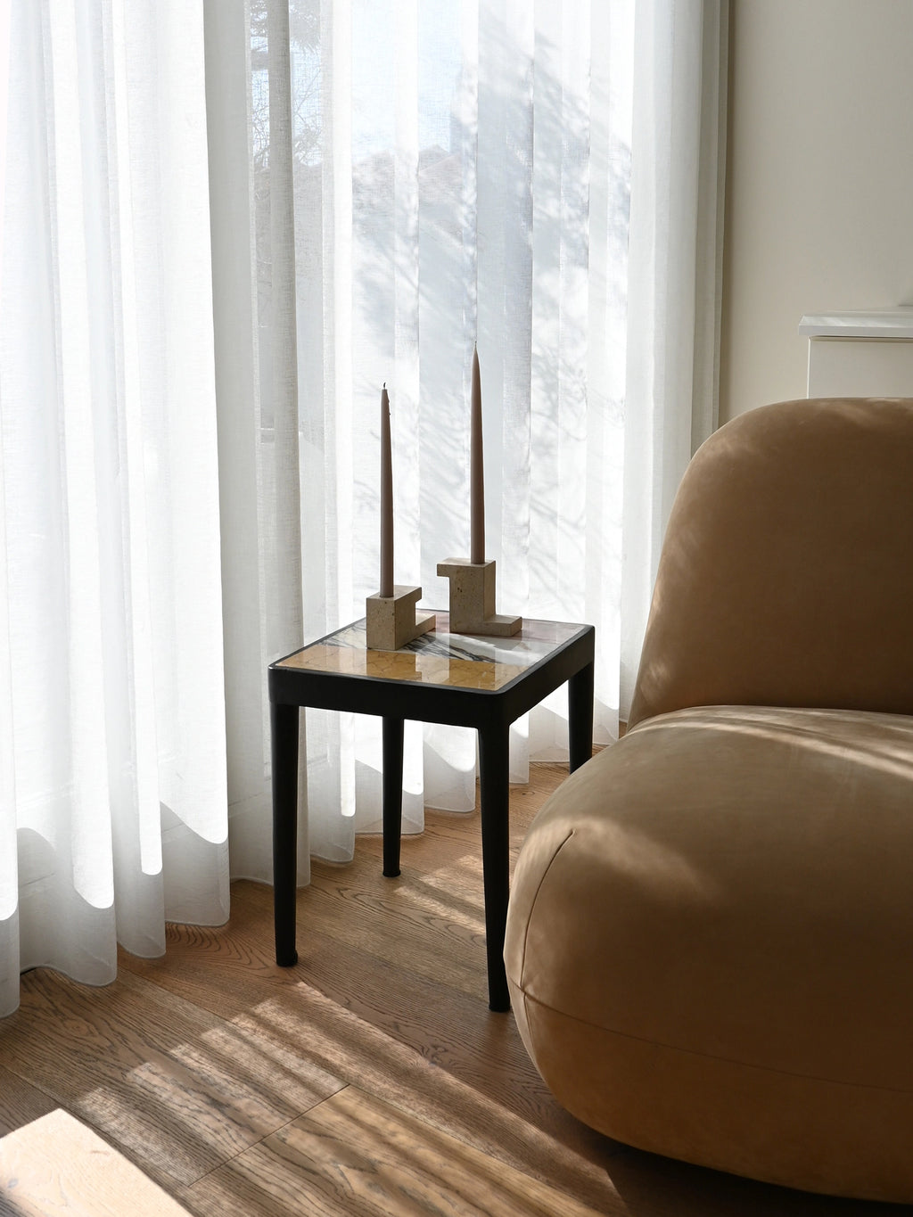 101 CPH Tairu Table: A modern black wooden side table with a square multicolored marble top, styled next to a beige velvet armchair, featuring two brown tapered candles. A premium accent for any elegant living space.