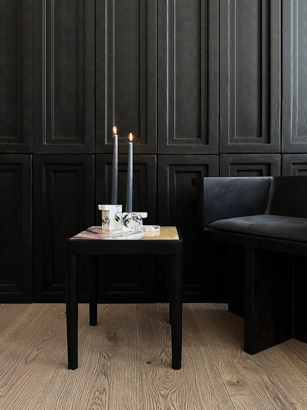 101 CPH Tairu Table, a luxurious black wooden side table with a square multicolored marble top, displayed in a modern dark room with a black bench and candles. Ideal for sophisticated living spaces.