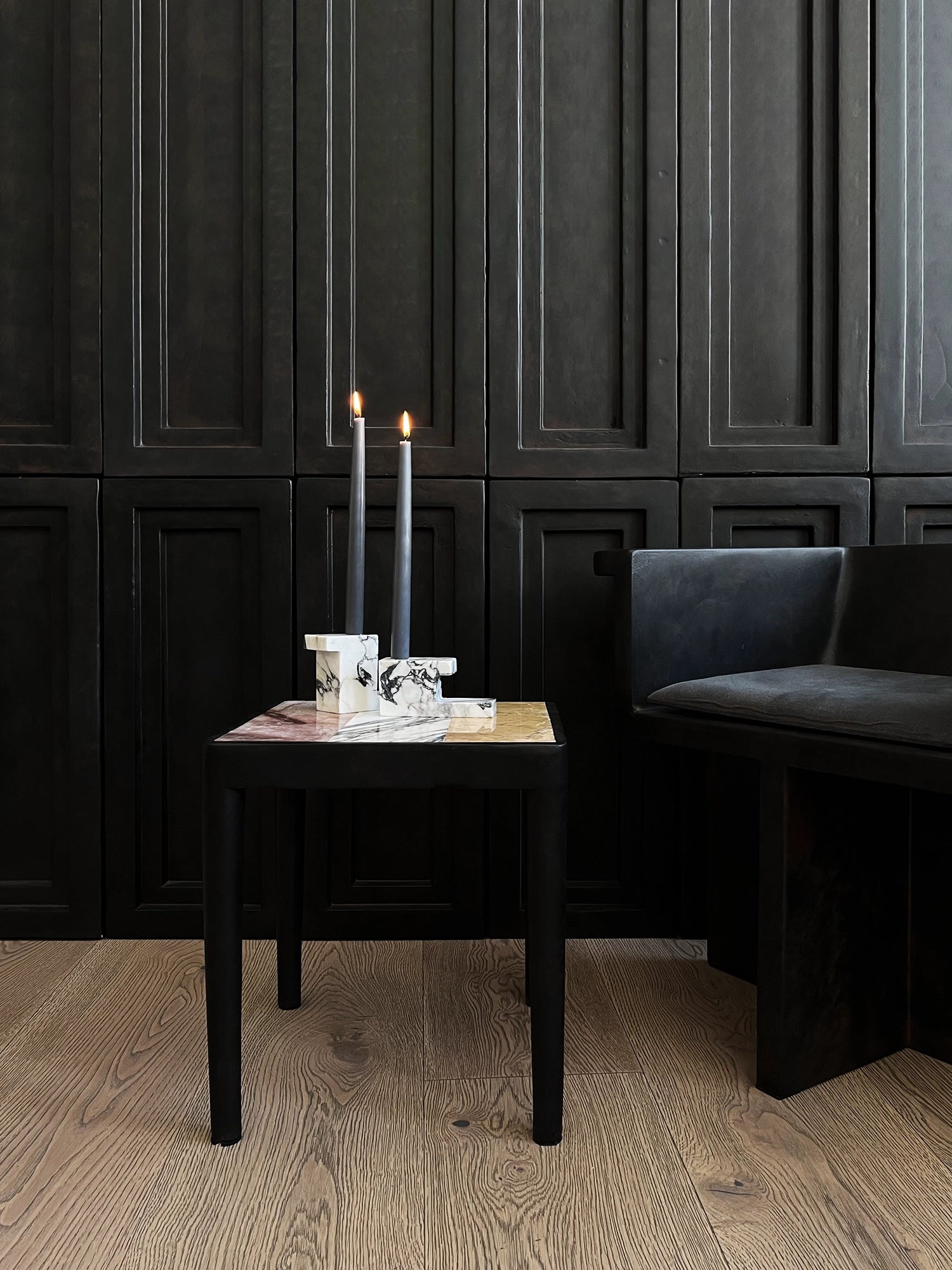 101 CPH Tairu Table, a luxurious black wooden side table with a square multicolored marble top, displayed in a modern dark room with a black bench and candles. Ideal for sophisticated living spaces.