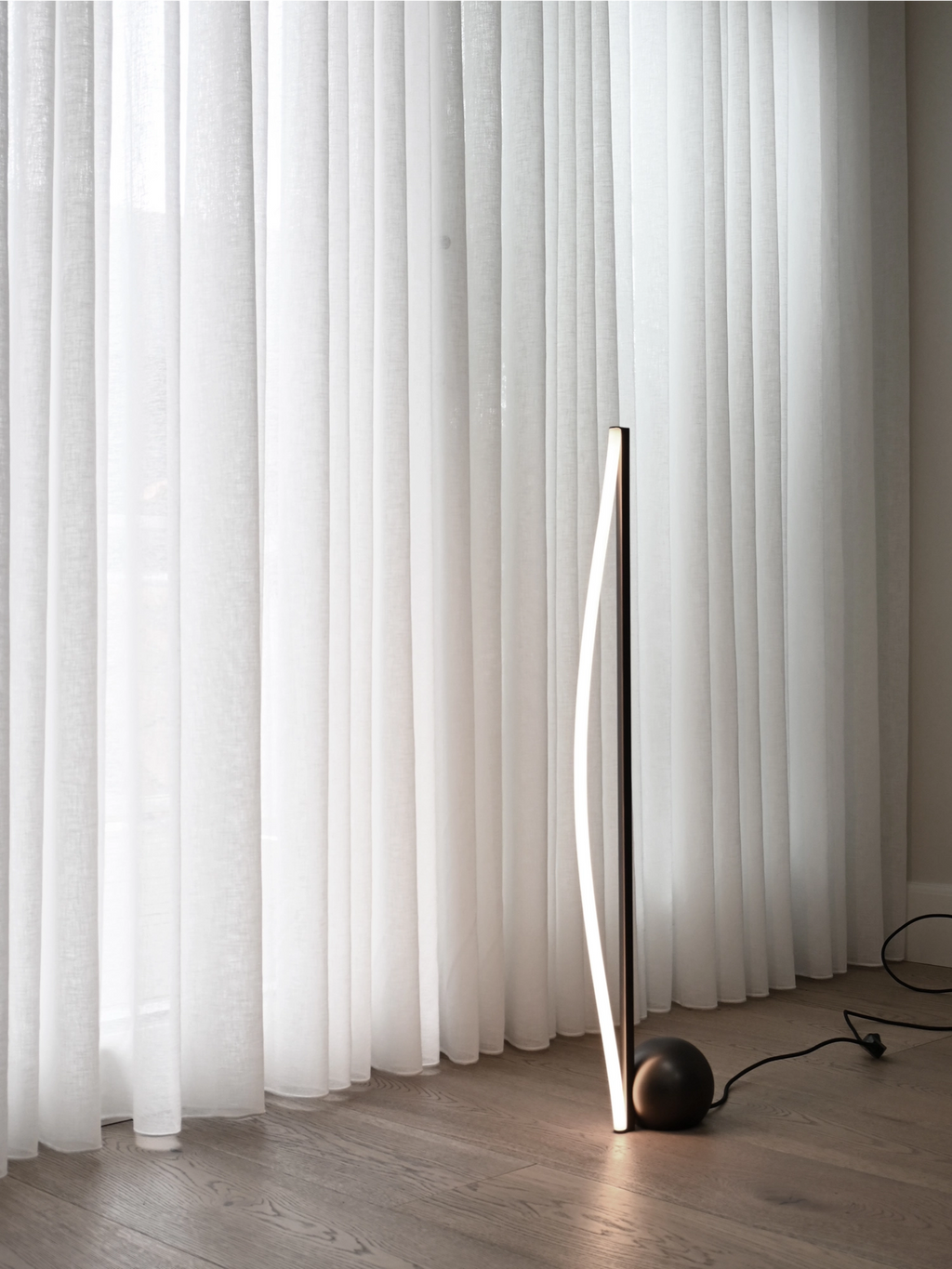 101 CPH Bow Floor Lamp, a modern, curved black metal LED lamp with a spherical base and glowing strip, set on a light wood floor beside white sheer curtains. A premium lighting solution.