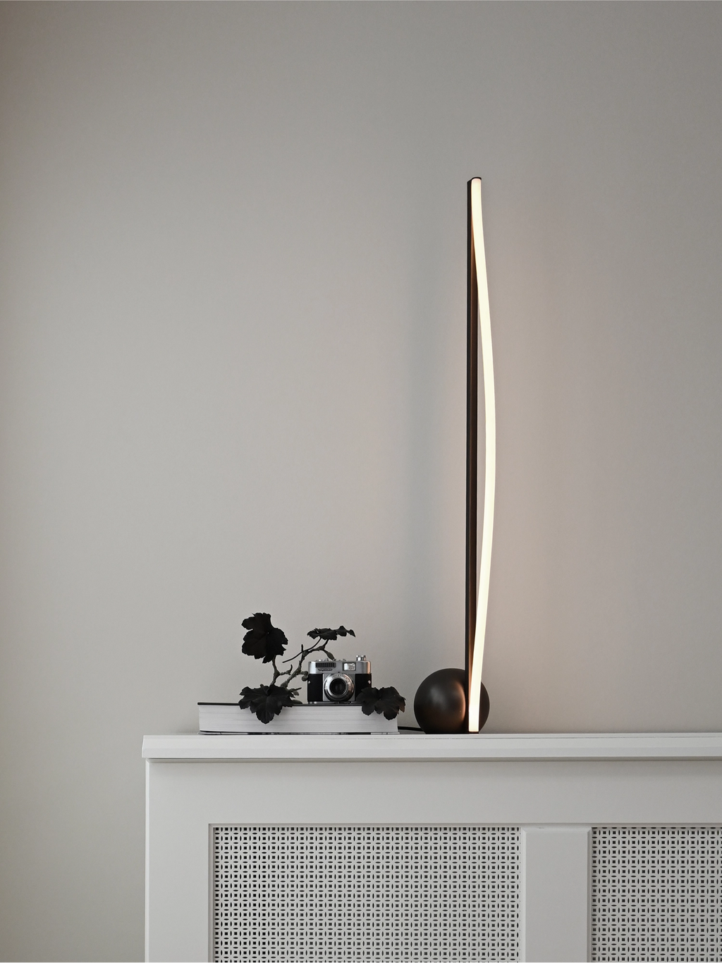 101 CPH Bow Floor Lamp, a modern matte black lamp with an illuminated, gently curved LED strip, on a white patterned radiator cover. Luxury lighting for contemporary spaces.