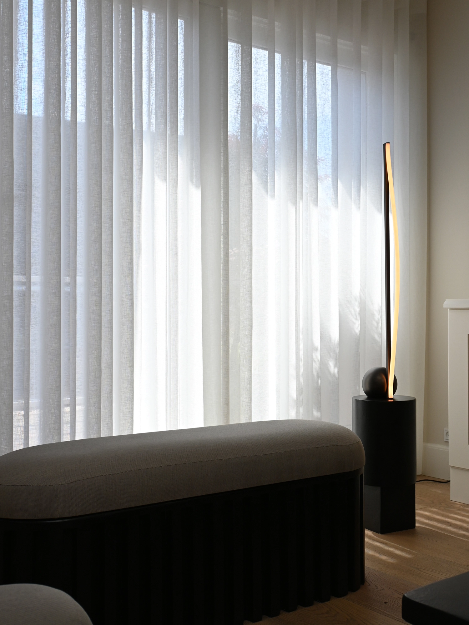 101 CPH Bow Floor Lamp, a modern sculptural design with a black circular base supporting an elegant curved light strip emitting warm amber light, in a luxurious living room setting. Ideal for contemporary interior illumination.