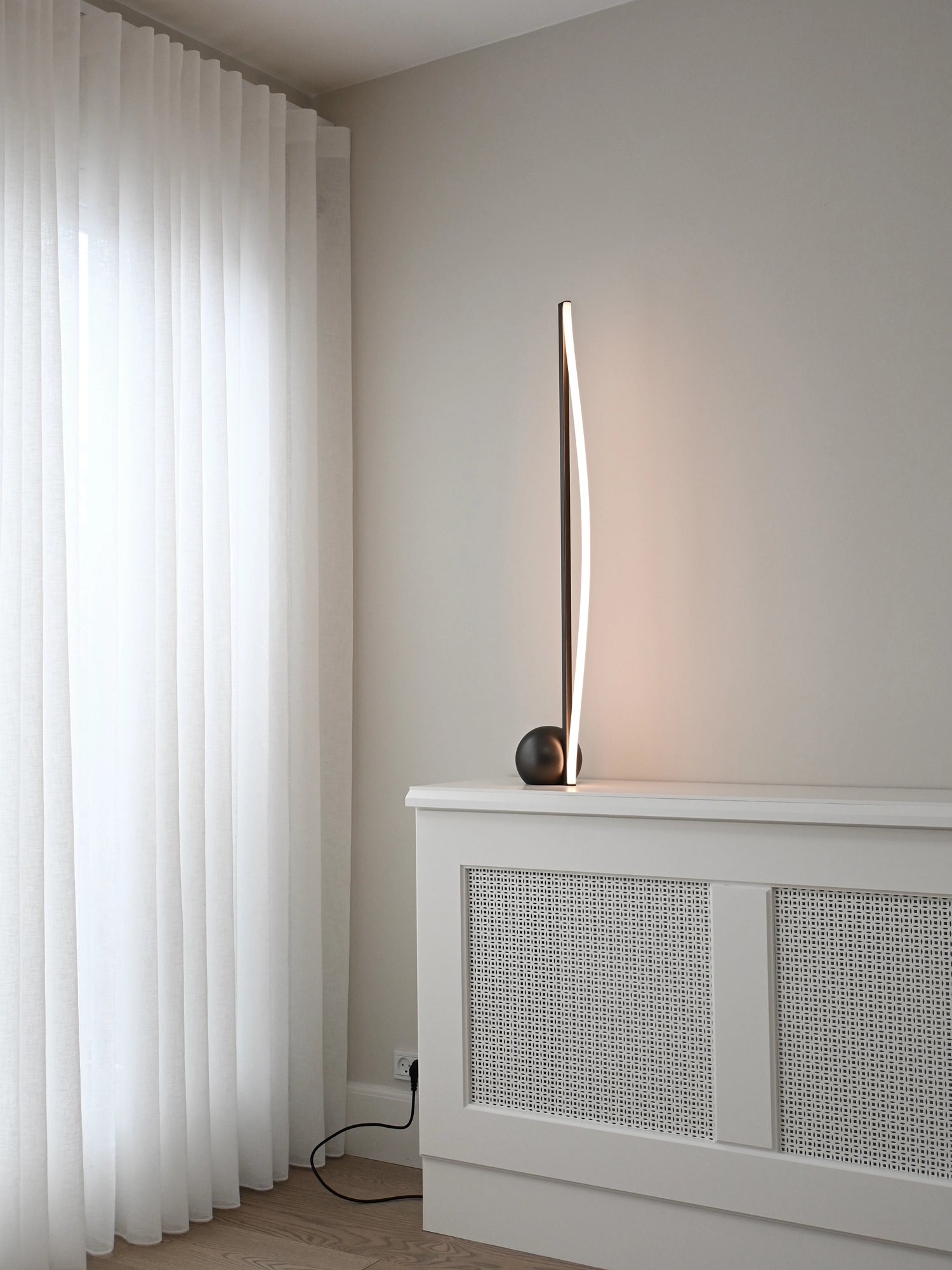 101 CPH Bow Floor Lamp, a modern matte black lamp with a curved LED strip emitting warm light, on a white radiator cover in a contemporary room. Ideal for minimalist aesthetic lighting.