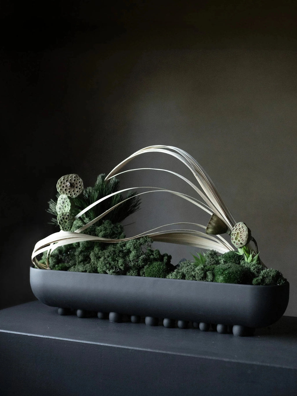 101 CPH Shinju Tray, a sleek matte black oblong tray with distinctive spherical feet, styled with lush green preserved moss, natural dried lotus pods, and delicate pale dried palm fronds for elegant organic decor.