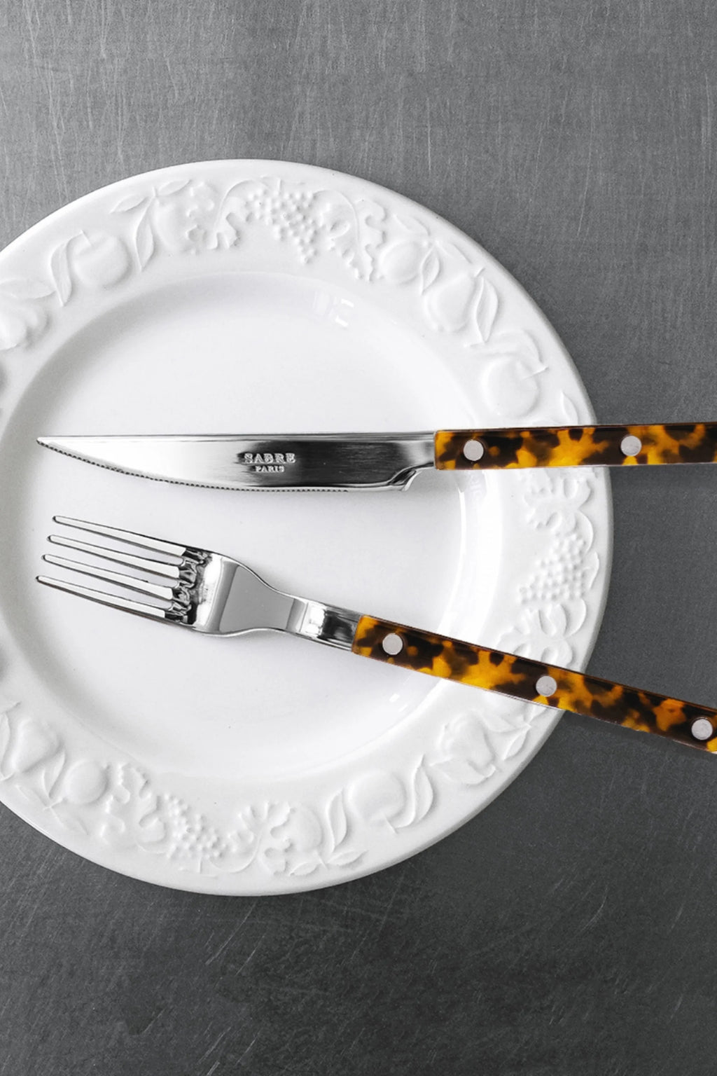 Sabre Paris Bistrot Tortoise 5-Piece Cutlery Flatware Set, featuring polished stainless steel with stylish tortoise shell effect resin handles and silver rivets, resting on a white embossed plate. This elegant flatware set is perfect for sophisticated dining and entertaining.