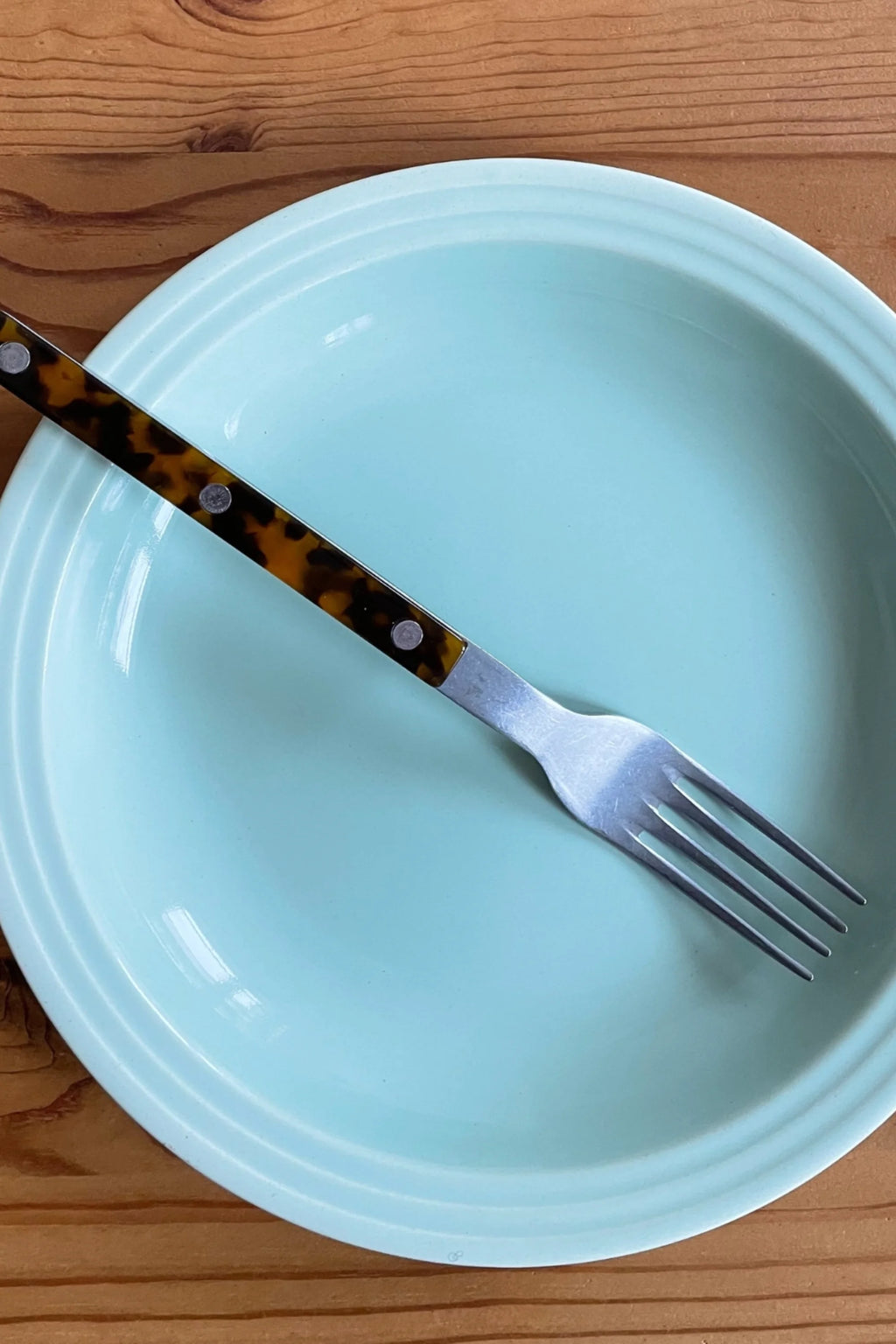 Sabre Paris Bistrot Vintage Tortoise 5-Piece Cutlery Flatware Set, featuring a polished stainless steel fork with a distinctive dark tortoise shell handle, accented with silver rivets, resting on a light blue plate atop a warm wooden table. This stylish flatware set is perfect for enhancing elegant dining experiences for those who appreciate vintage-inspired kitchenware.