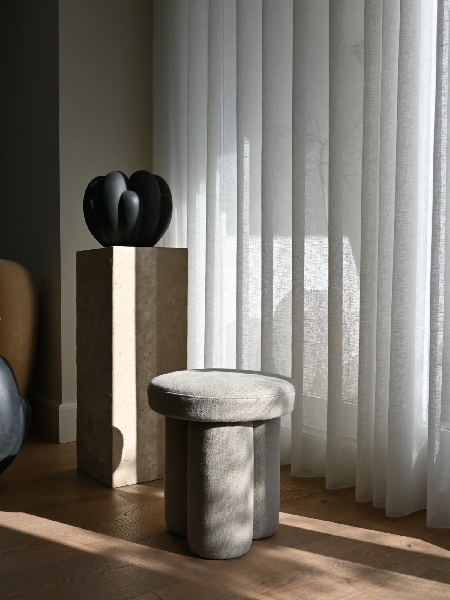 101 CPH Big Foot Stool, stylish modern light gray fabric stool with three sturdy, rounded legs and a soft, padded top, casting elegant shadows on a dark wooden floor. Perfect for adding a touch of minimalist Scandinavian design and comfort to a contemporary living room, bedroom, or entryway for design-conscious homeowners.