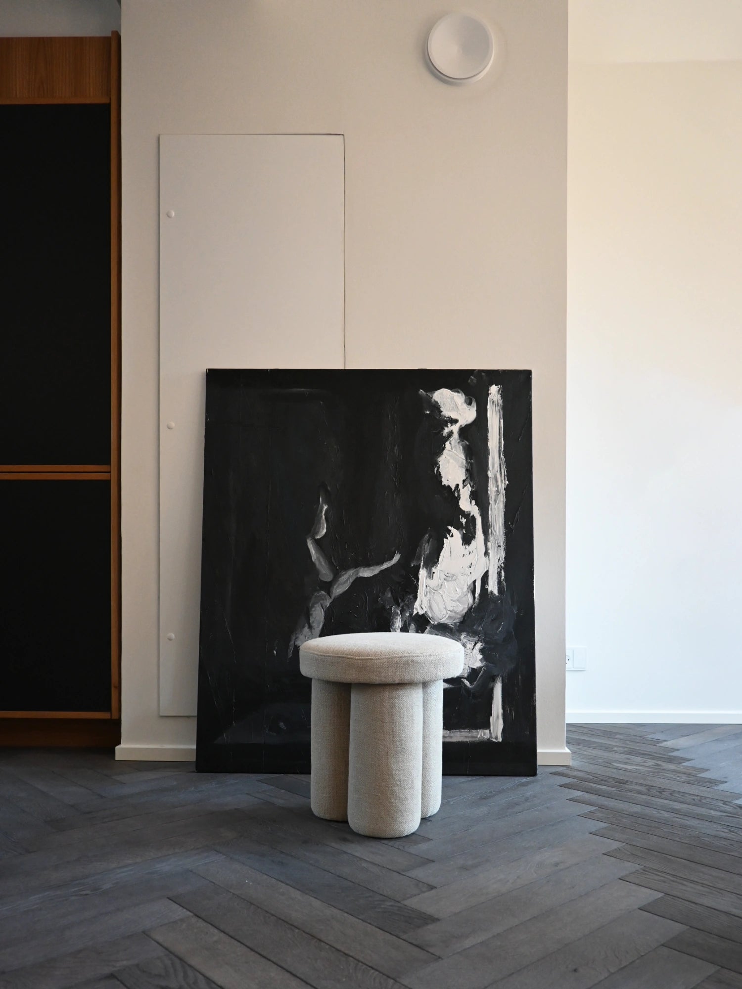 101 CPH Big Foot Stool, elegant cream-colored fabric stool with a thick round top and three stout, cylindrical legs standing on dark grey herringbone wood flooring. This modern, sculptural piece adds stylish, functional seating or a unique accent to any contemporary living space or bedroom.