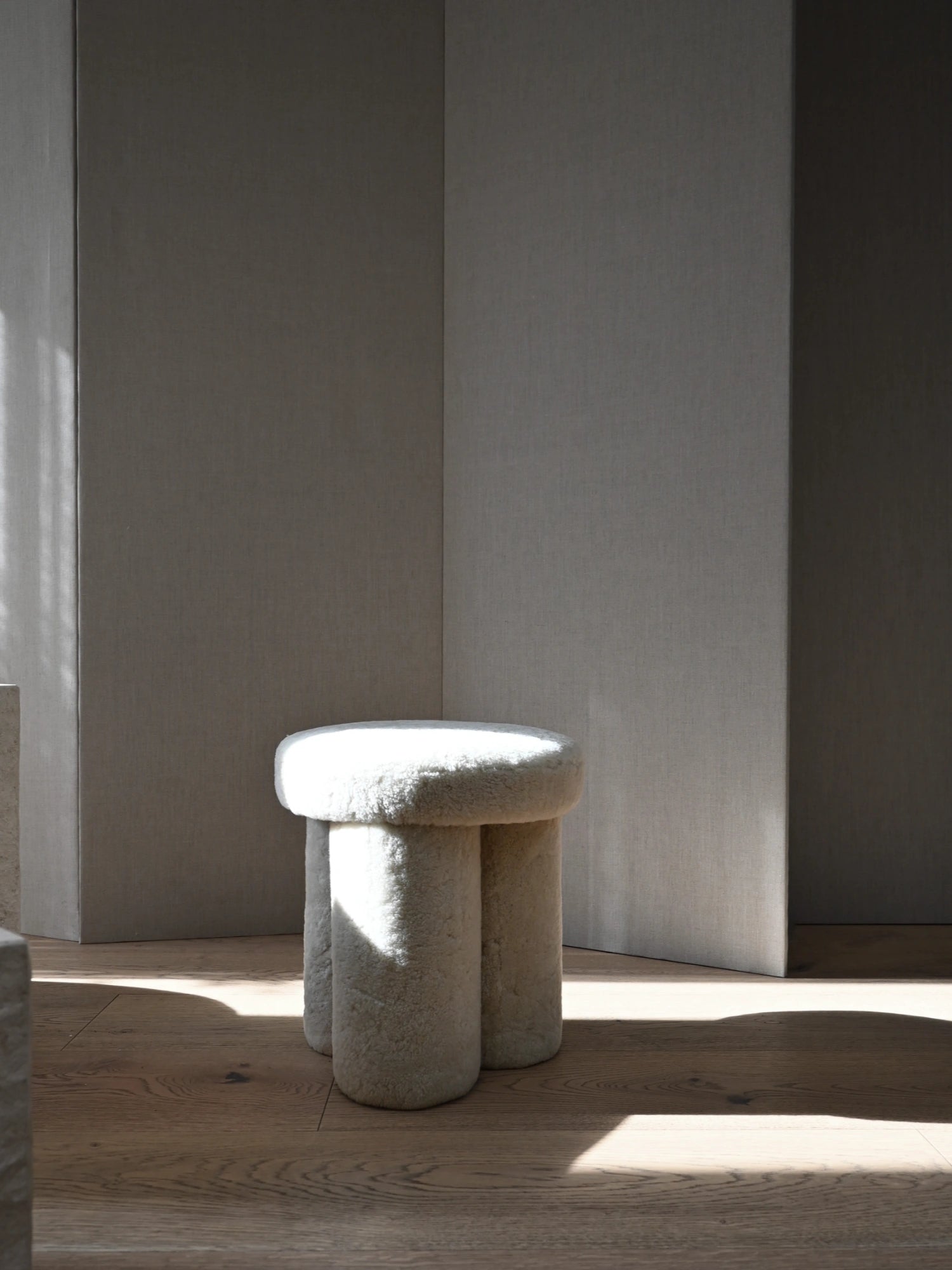 101 CPH Big Foot Stool, elegant cream-white sheepskin stool with three thick, sculptural legs and a plush rounded top on warm oak hardwood flooring. This luxurious, modern stool is perfect as a stylish accent piece or comfortable seating for contemporary interior design enthusiasts.