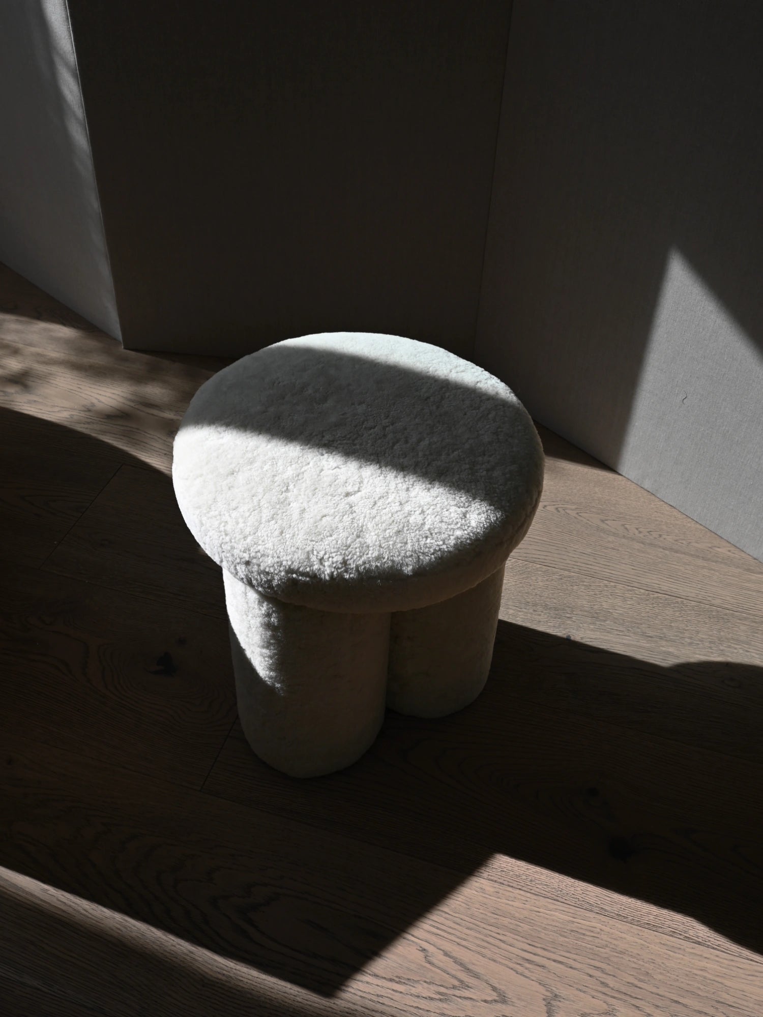 101 CPH Big Foot Stool, stylish plush off-white bouclé texture stool with three rounded legs on a warm wooden floor, accented by natural light and shadows. A luxurious modern seating option for contemporary living spaces or cozy reading nooks.