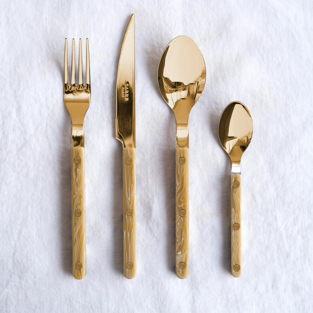 Sabre Paris Bistrot Horn Cutlery Set, featuring glossy gold-toned stainless steel utensils with elegant, swirled beige and white resin handles secured by brass rivets. This luxurious flatware set is perfect for enhancing sophisticated dinner parties and everyday dining for discerning hosts.