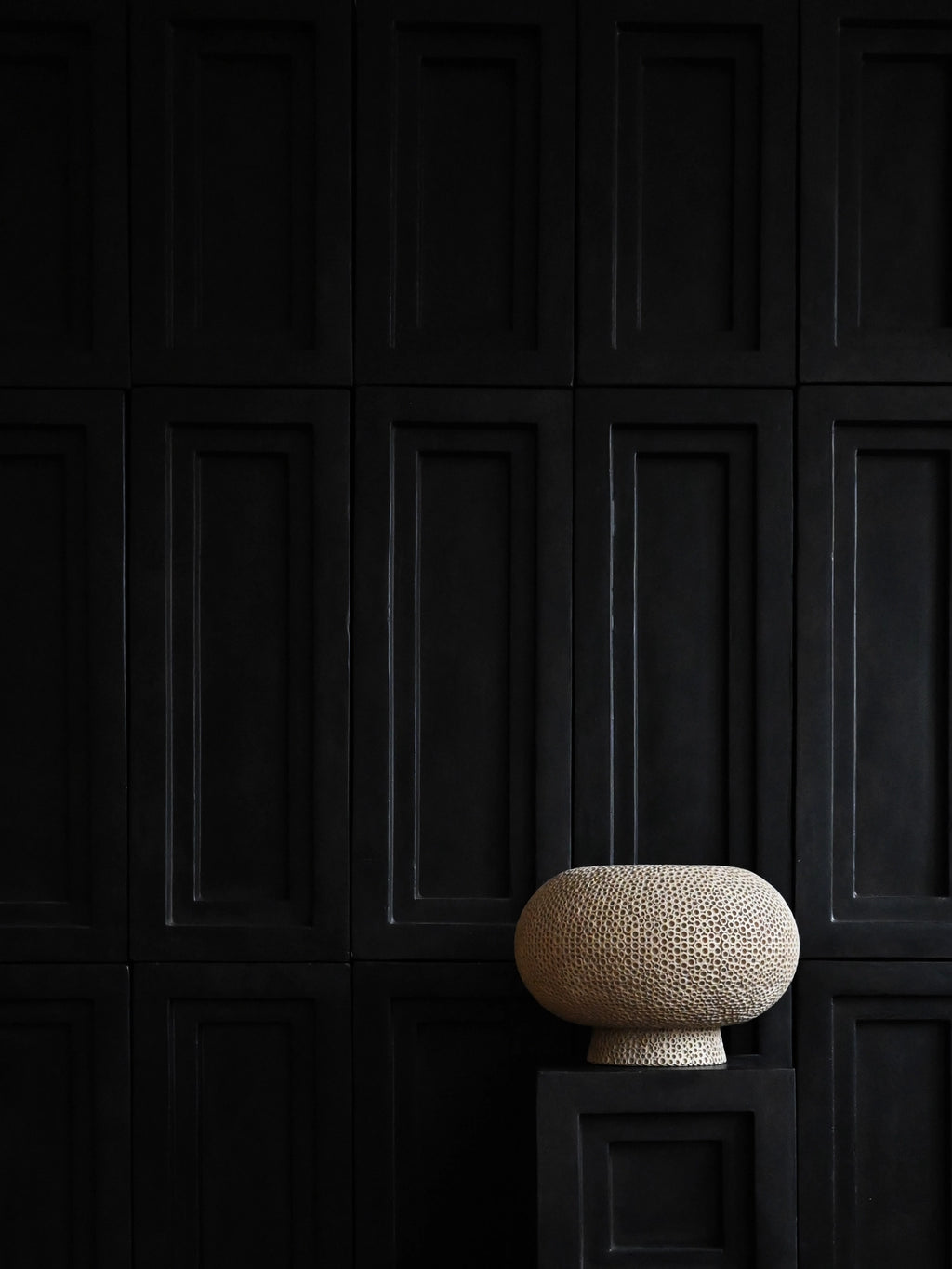 101 CPH Kabin Vase - Shisen: Oval-shaped, handcrafted beige ceramic decorative vase with a textured, porous surface and small pedestal base, displayed on a black geometric stand against a dark paneled wall. Perfect for unique home decor.