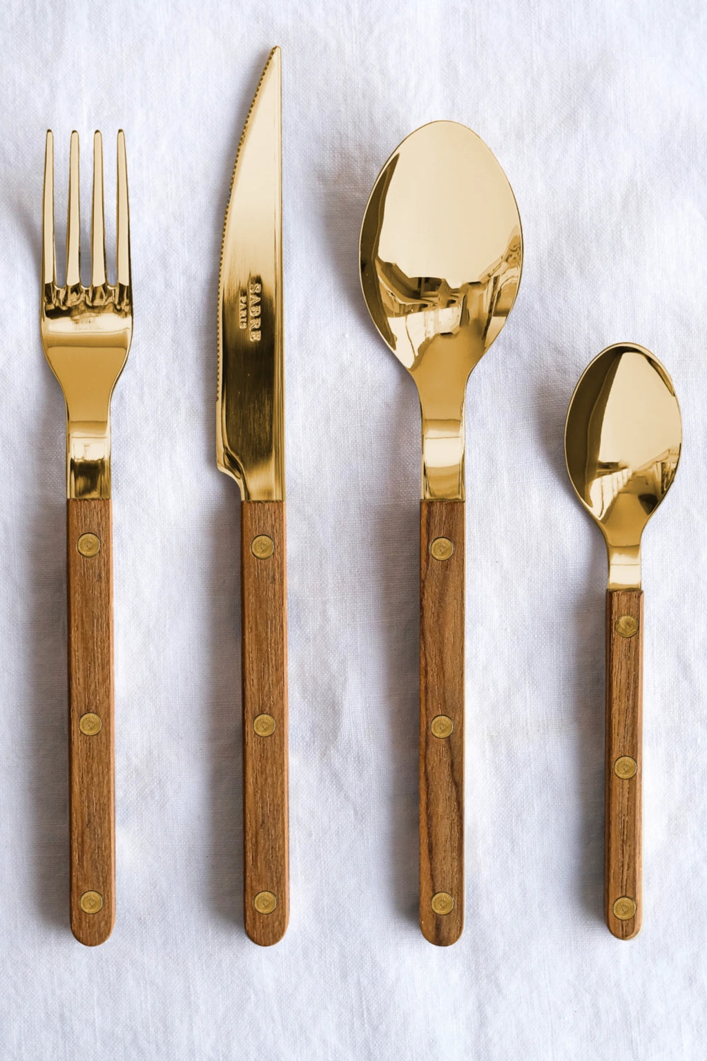 Sabre Paris Bistrot Gold Teak 5-Piece Cutlery Flatware Set, featuring polished gold-tone stainless steel with warm, natural teak wood handles and brass rivets, arranged neatly on a white linen tablecloth. This luxury flatware is perfect for elevating everyday dining or special occasions for those who appreciate elegant home decor.