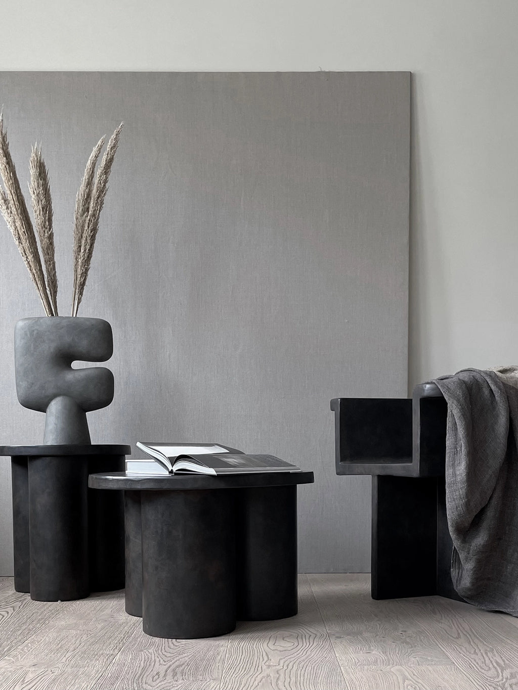 101 CPH Big Foot Table. Two matte dark grey, textured concrete tables with thick cylindrical legs, featuring an open book and abstract vase with dried pampas grass. A luxury decor statement.