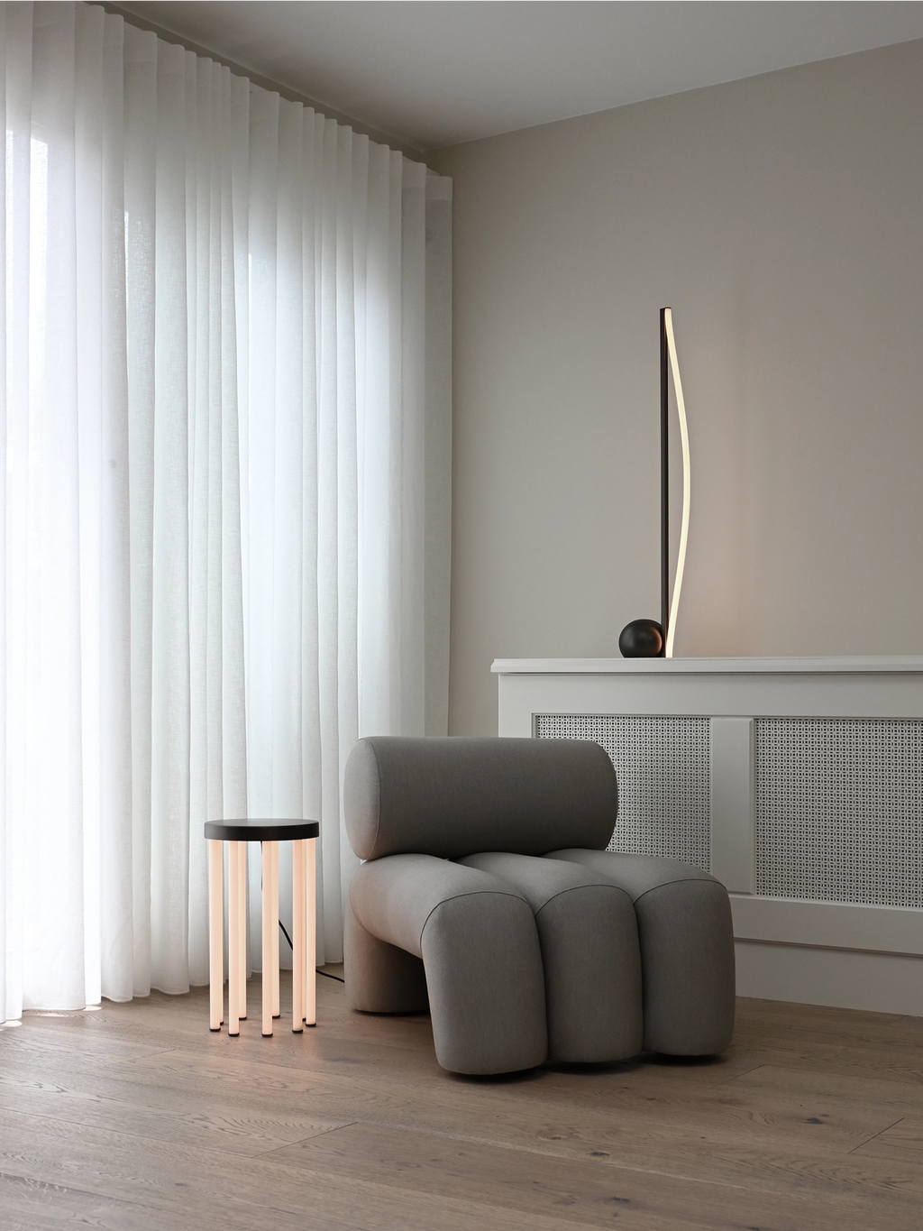 101 CPH Neon Side Table featuring a round black top and illuminated tubular legs, casting a soft modern glow on a wooden floor, next to a grey lounge chair. A minimalist luxury statement for any contemporary living space.