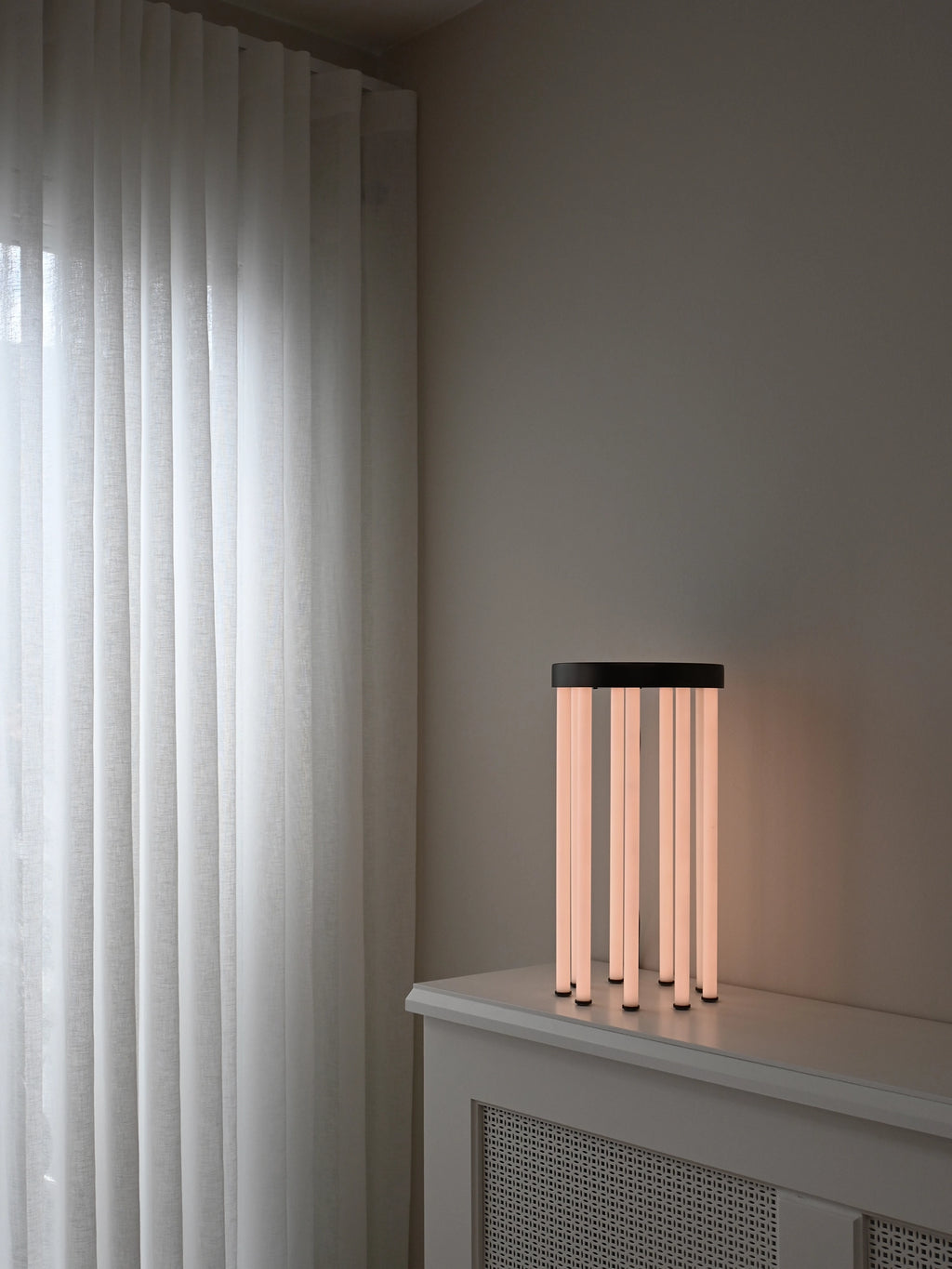 101 CPH Neon Side Table featuring multiple glowing, vertical cylindrical lamps with a dark circular top, creating a warm diffused light. This luxury modern lighting piece rests on a white textured cabinet, next to sheer white curtains. Ideal for contemporary interior design.