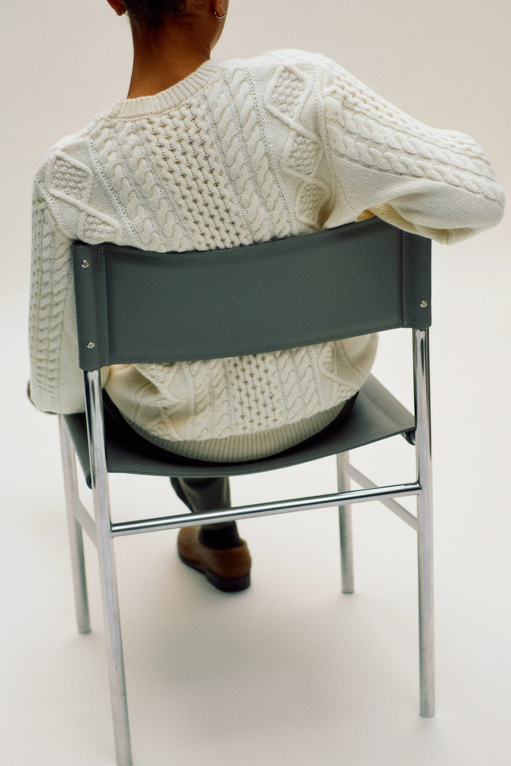 Person wearing a white cable knit sweater sitting on a black chair against a white background
