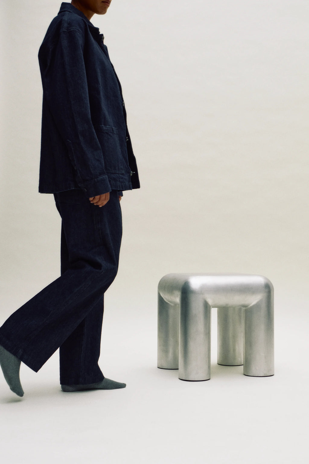Person wearing a dark outfit standing next to a metallic stool on a plain background