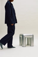 Person wearing a dark outfit standing next to a metallic stool on a plain background